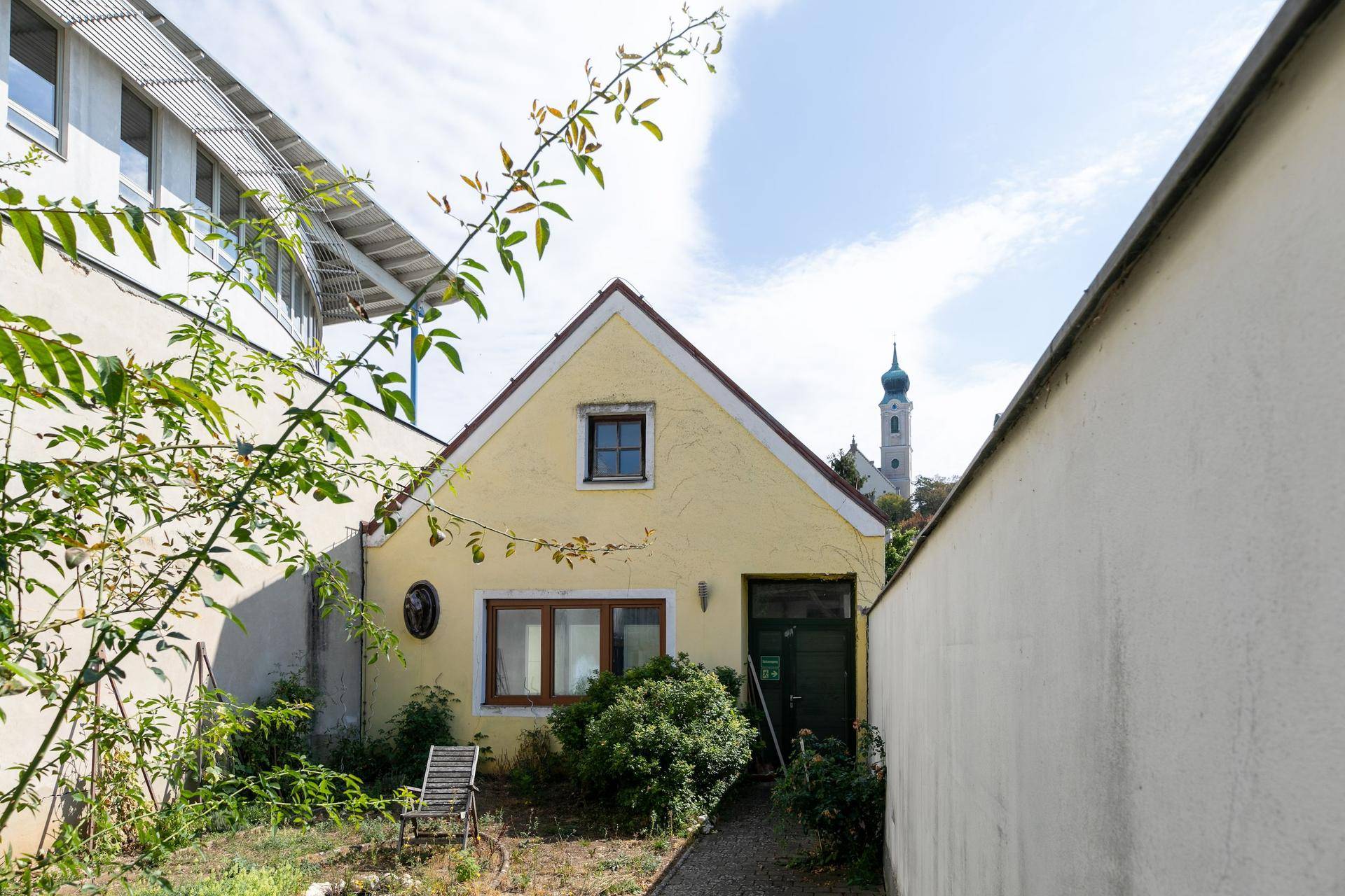 Kleines gelbes Haus mit Satteldach und einem schmalen Gartenbereich, im Hintergrund eine Kirche.