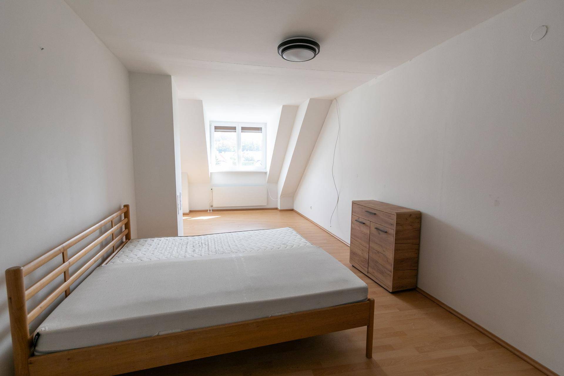 Schlafzimmer mit hellem Holzbett, Kommode, Parkettboden und Fenster in der Dachschräge.