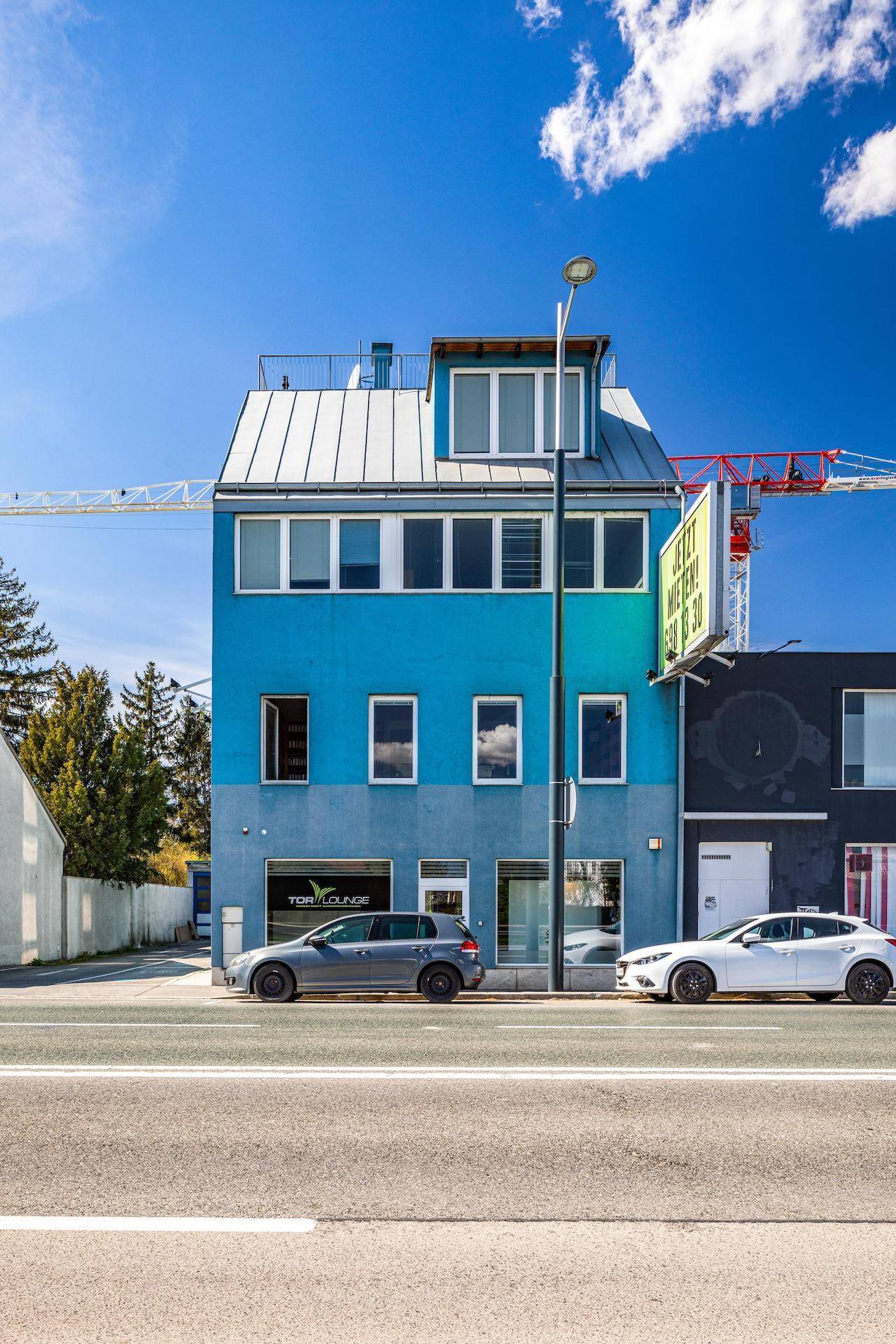 Modernes blaues Gebäude mit großen Fenstern und einem Flachdach, gelegen an einer städtischen Straße.