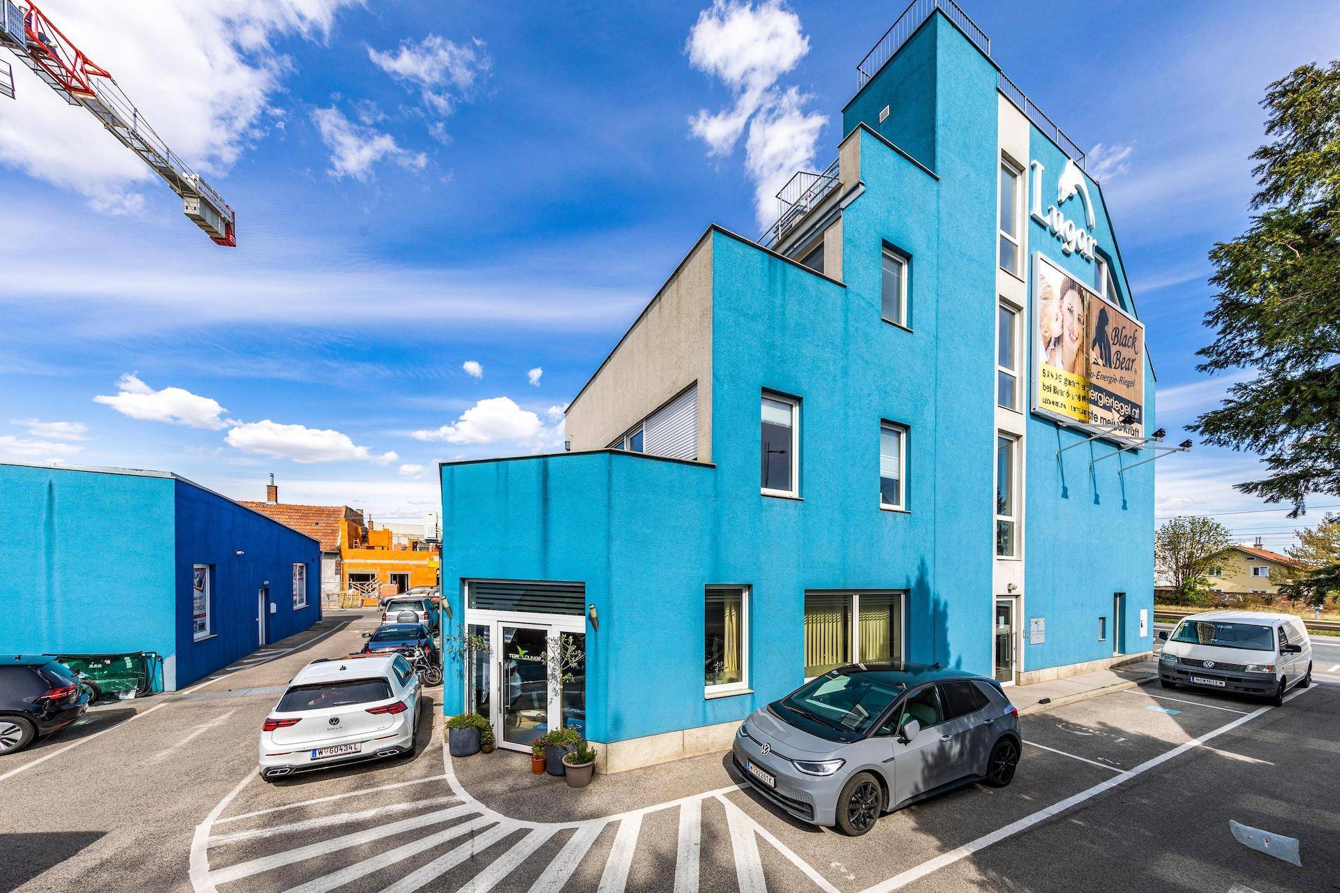 Modernes blaues Gebäude mit Parkplätzen davor und einem Baugerüst im Hintergrund unter blauem Himmel.
