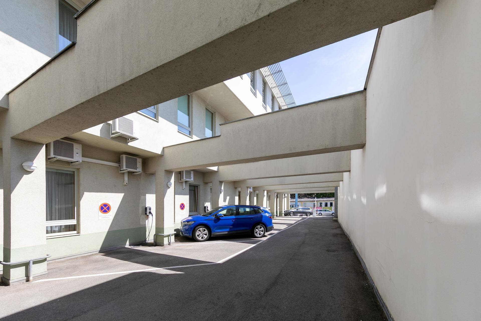 Überdachter Parkplatzbereich mit einem blauen Auto und Klimaanlagen an der Fassade.