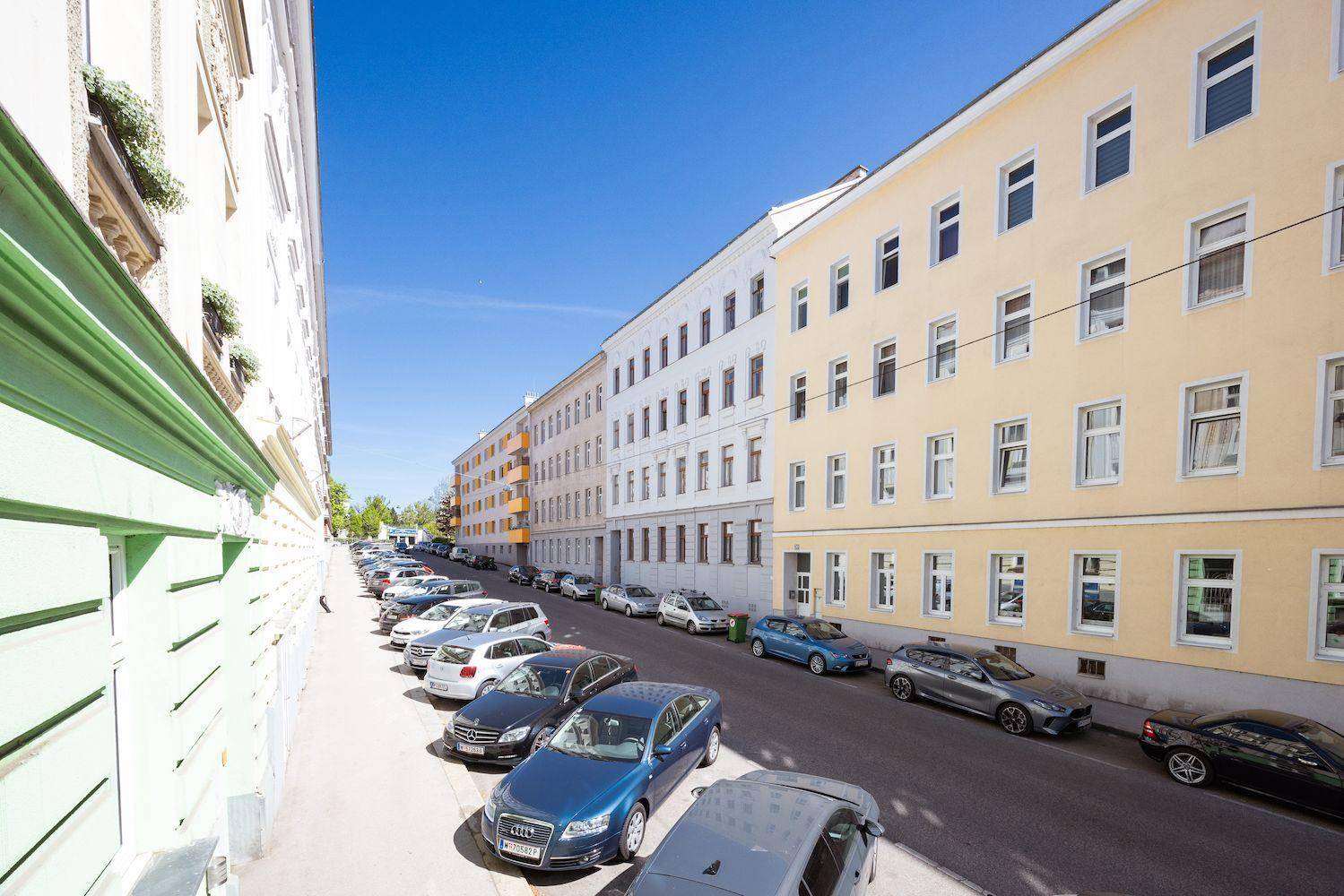 Blick entlang einer belebten Stadtstraße mit parkenden Autos und Gebäudereihen unter blauem Himmel.