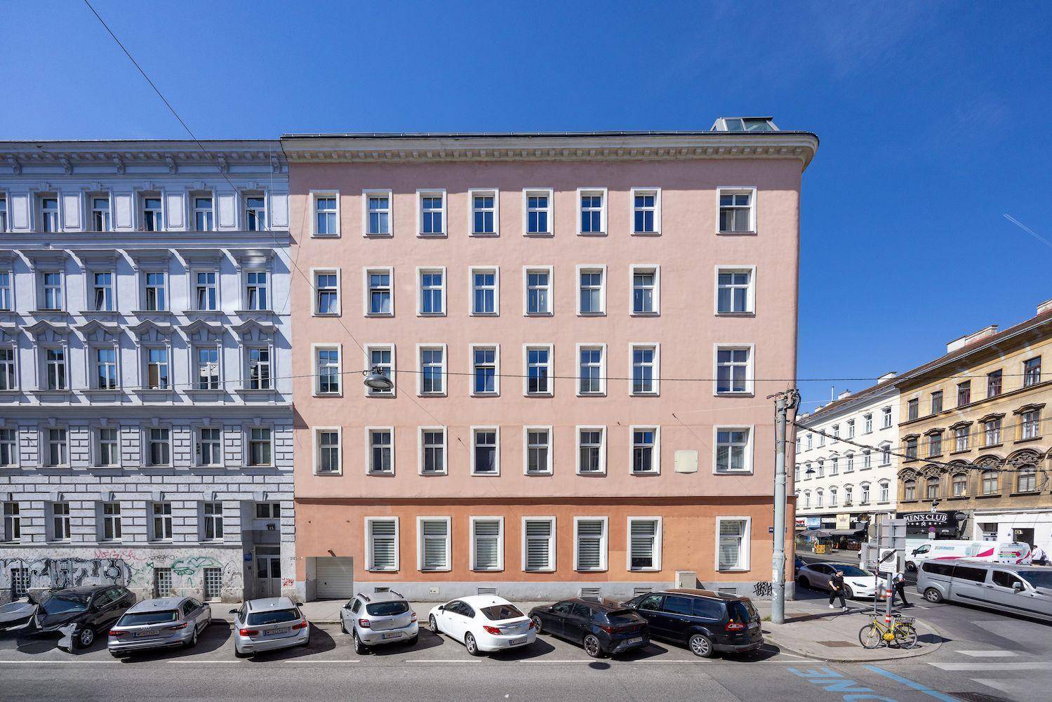 Frontale Ansicht eines historischen Gebäudes mit vielen Fenstern und geparkten Autos auf der Straße.