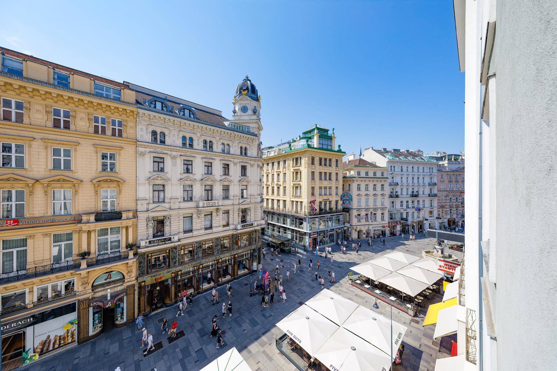 Belebter Stadtplatz mit historischen Gebäuden, Geschäften und Außengastronomie unter blauem Himmel.