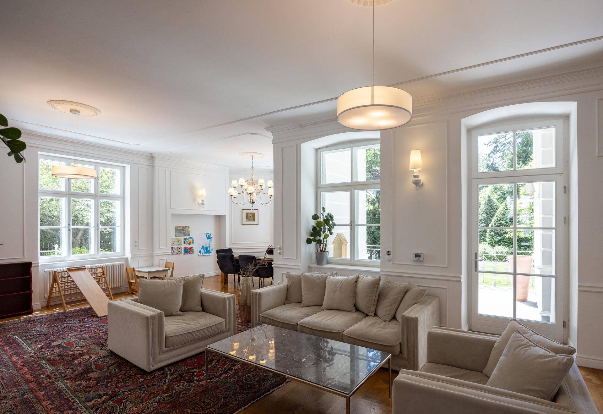 Großzügiges Wohnzimmer mit hohen Decken, Stuckelementen und Blick in den grünen Garten.