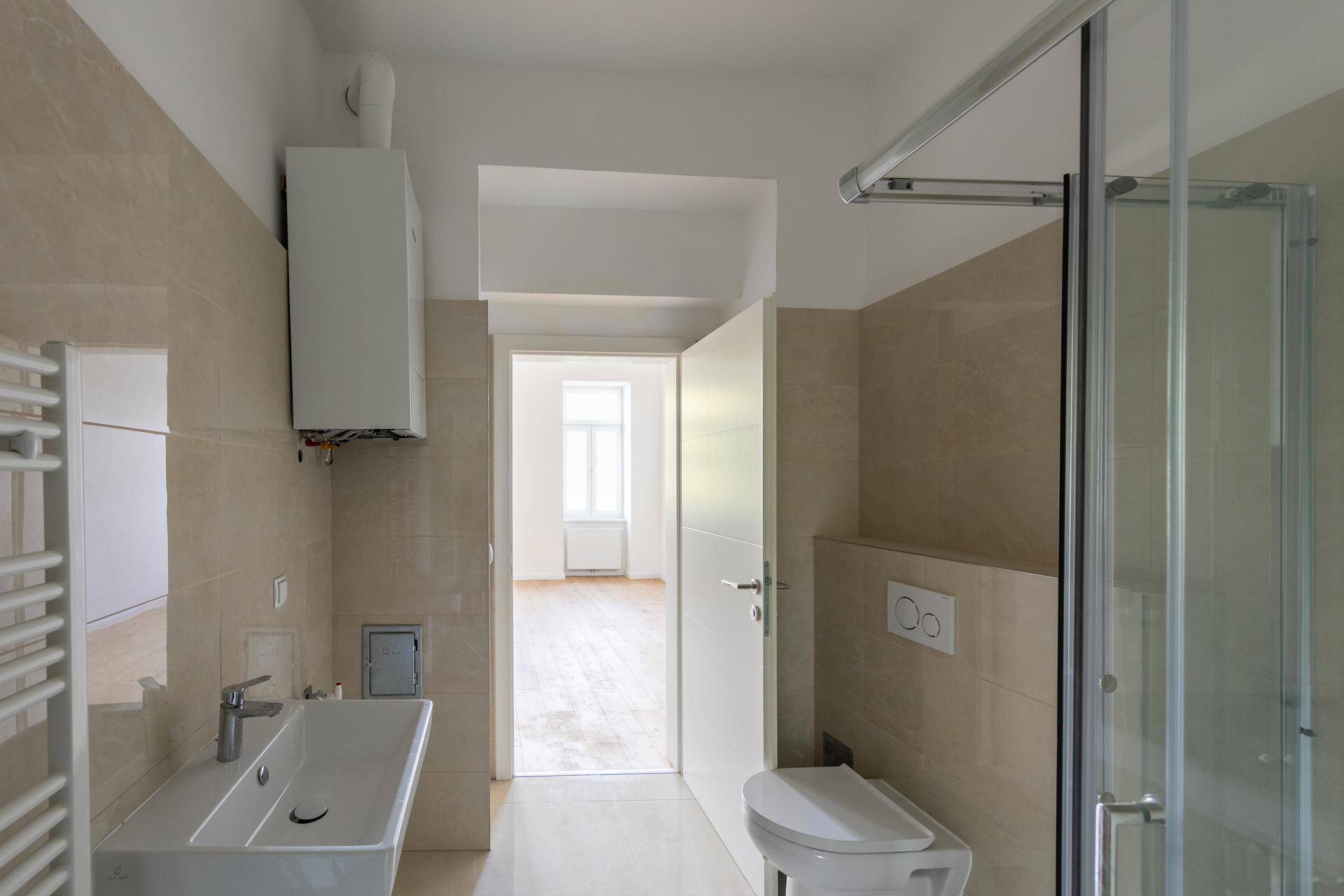 Helles Badezimmer mit Dusche, Waschbecken und Heizkörper, Blick in einen weiteren Raum.