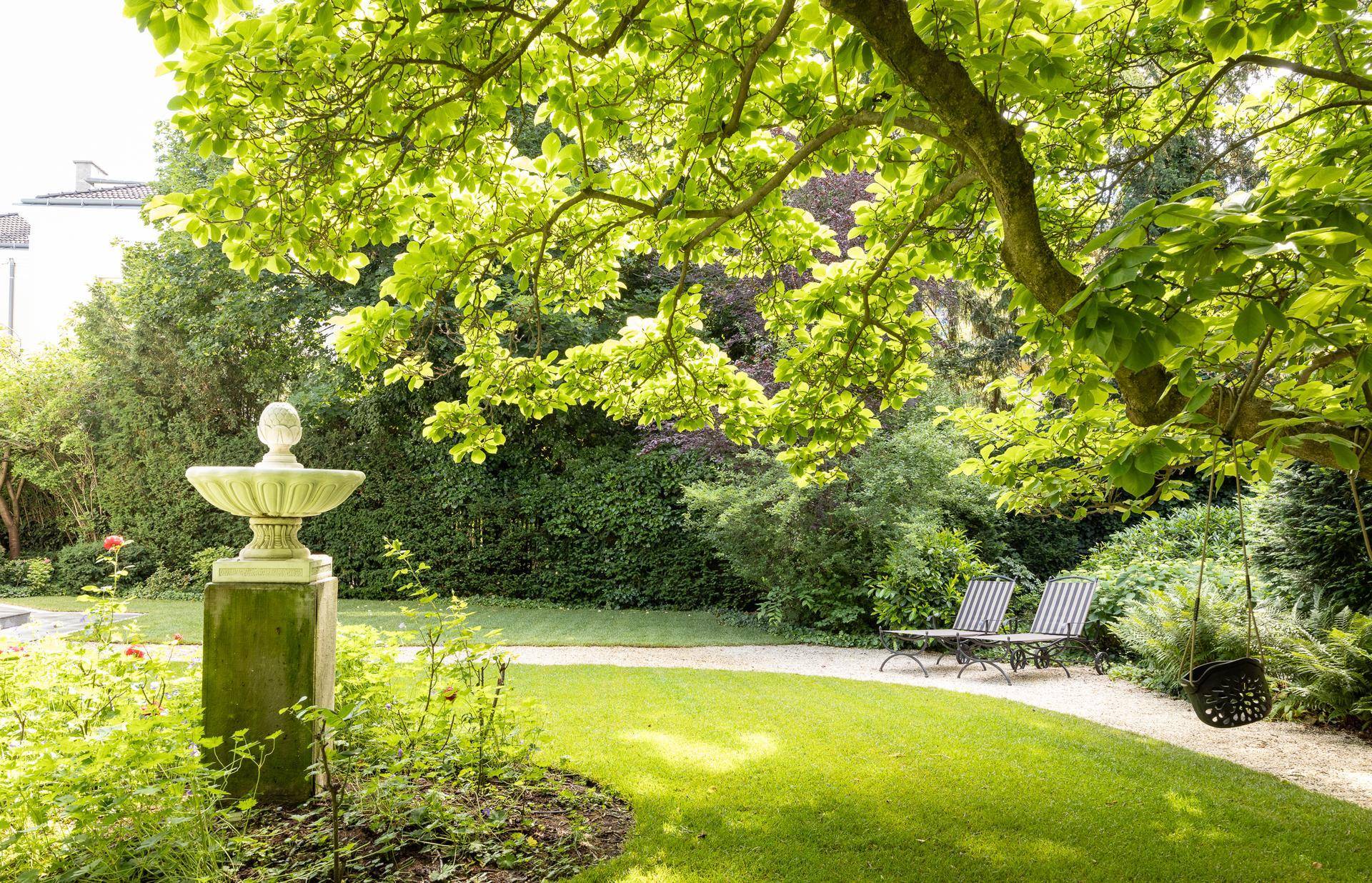 Ruhiger Gartenbereich mit dekorativem Brunnen, Liegestühlen und üppigem Grün.