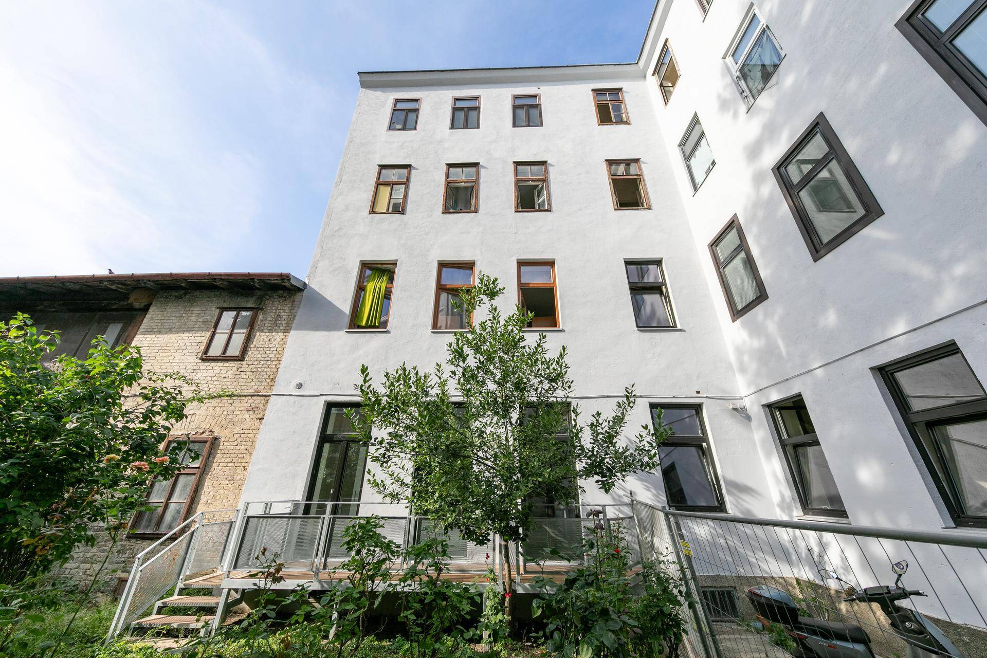 Rückansicht eines Mehrfamilienhauses mit mehreren Etagen und Bäumen im Vordergrund.