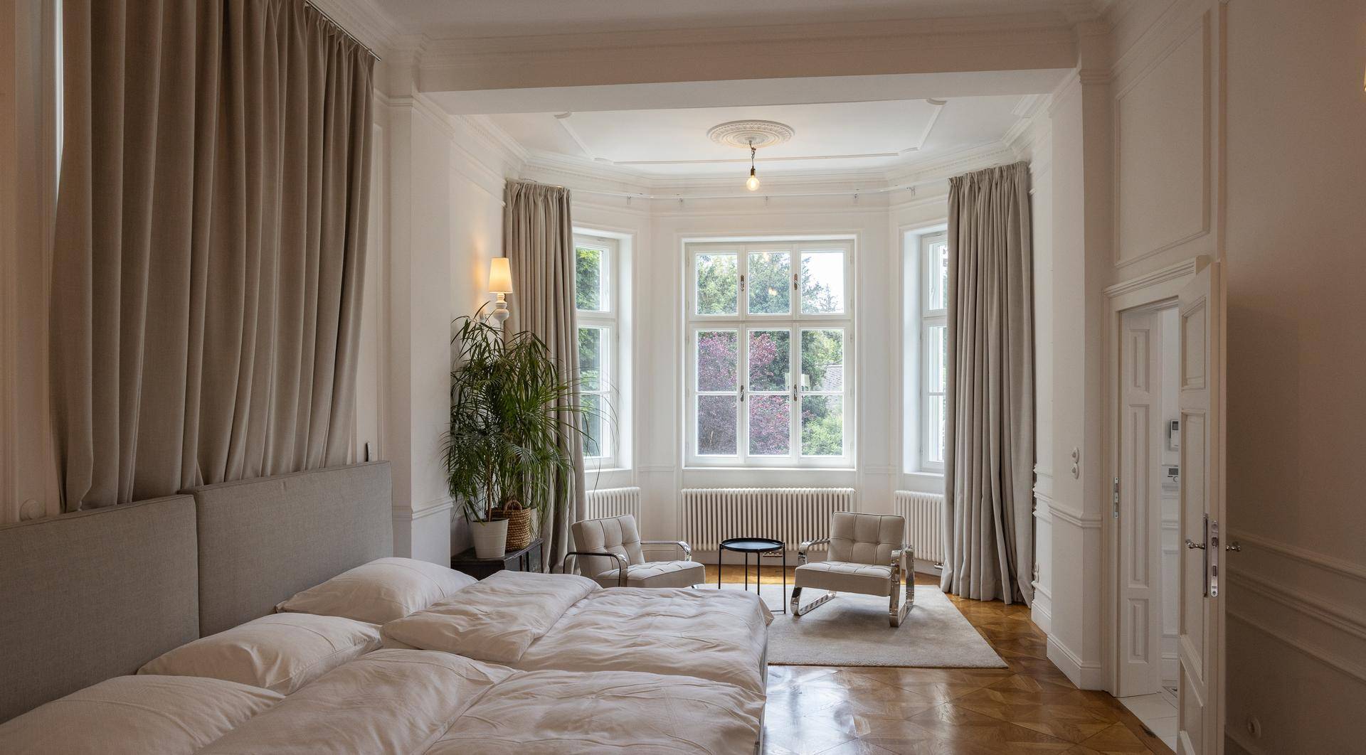 Elegantes Schlafzimmer mit großem Bett, hohen Decken und einer hellen Sitzecke im Erker.