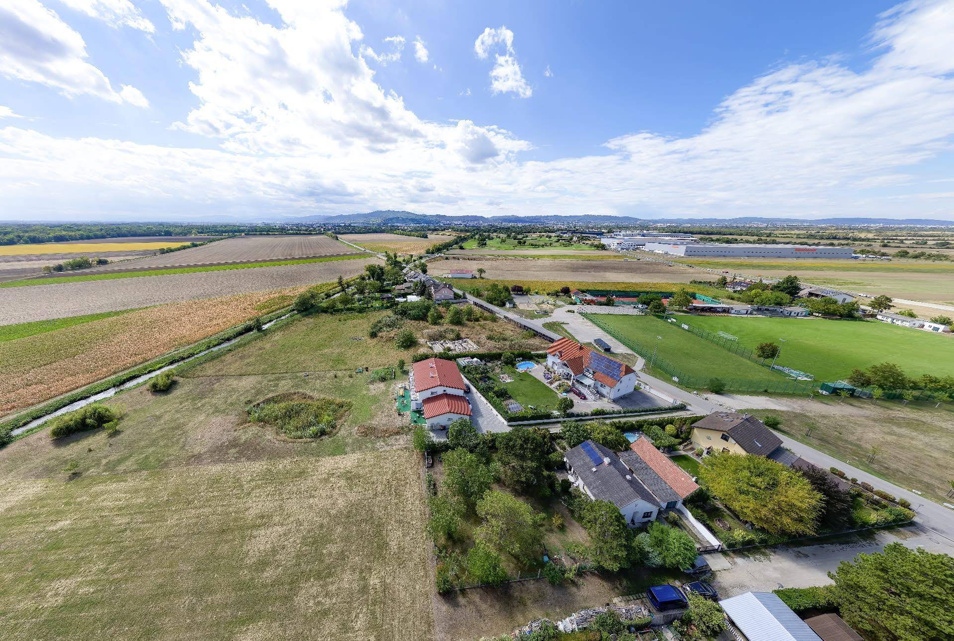 Panoramablick aus der Luft auf die ländliche Umgebung mit Feldern, Häusern und einer weiten Landschaft unter blauem Himmel.