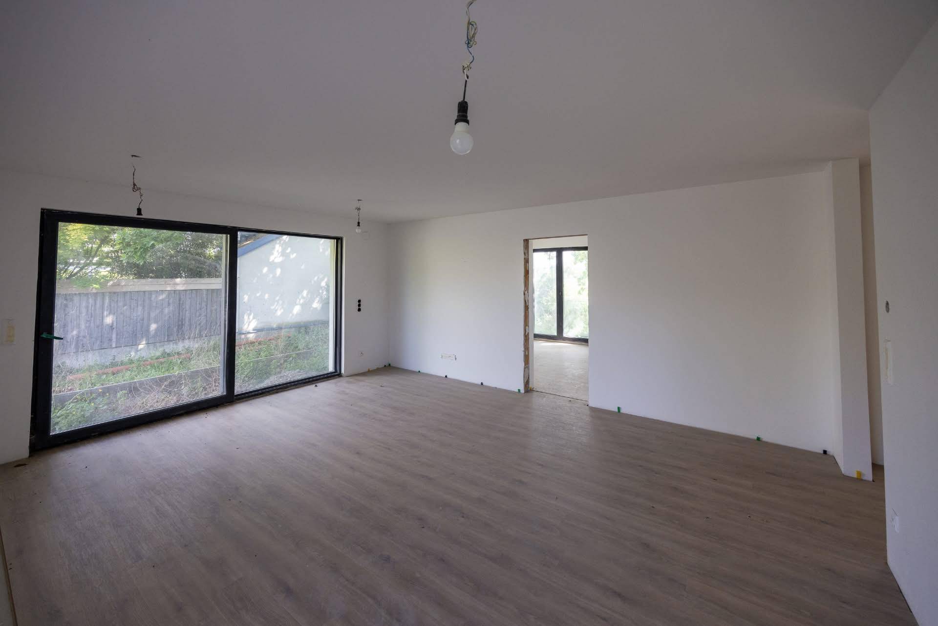 Geräumiger Wohnbereich im Rohbau mit großen Schiebetüren und neu verlegtem Laminatboden.