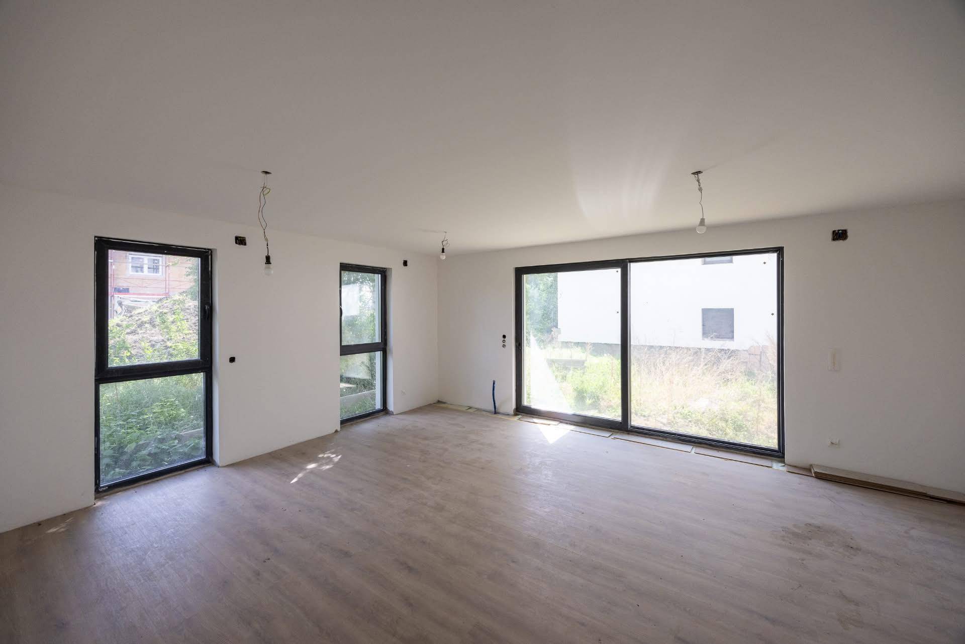 Heller Wohnbereich im Rohbau mit großen Fenstern und Schiebetüren, teilweise mit Laminatboden.
