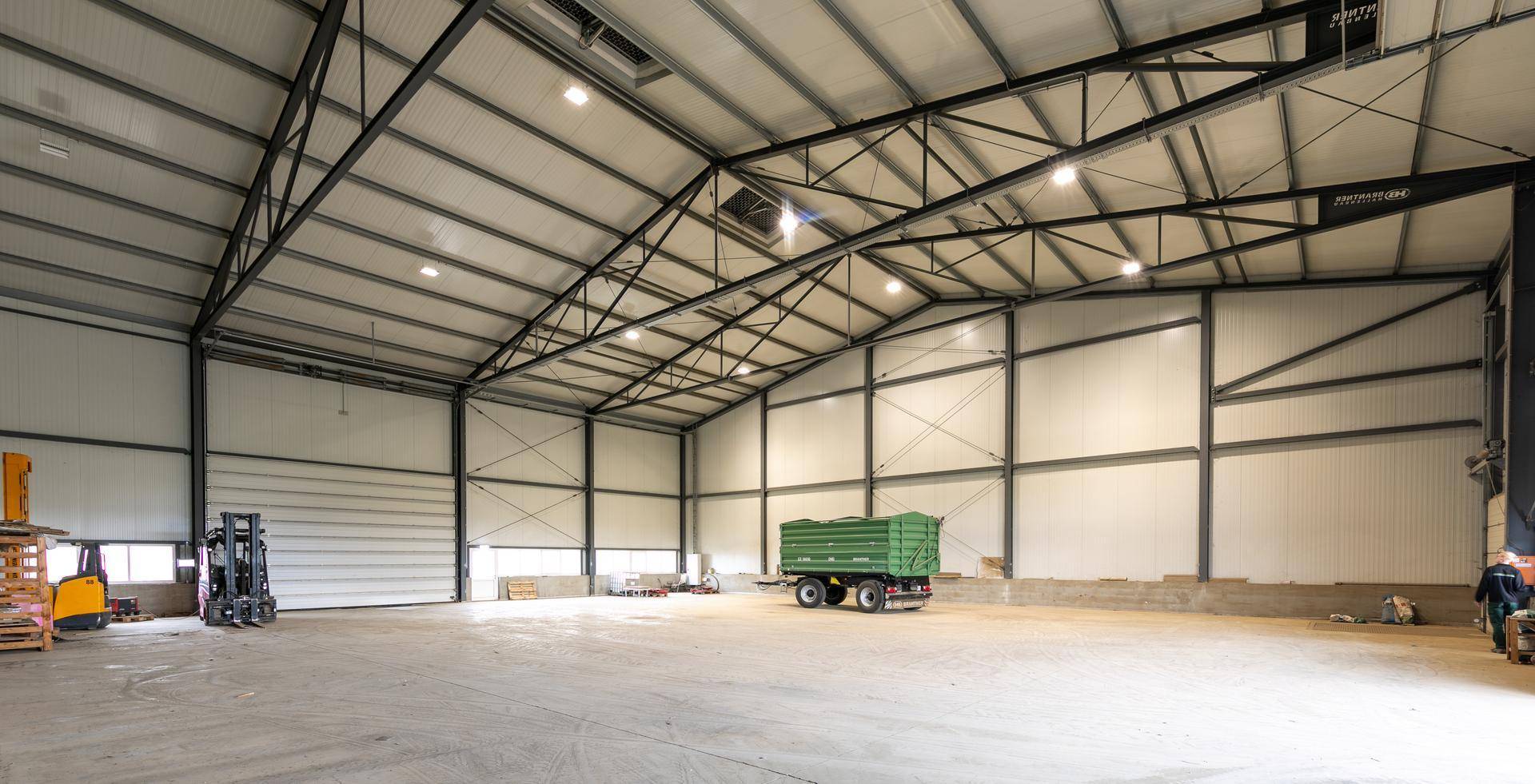 Geräumige Lagerhalle mit hohen Decken, Metallkonstruktion und einem landwirtschaftlichen Anhänger auf dem Betonboden.