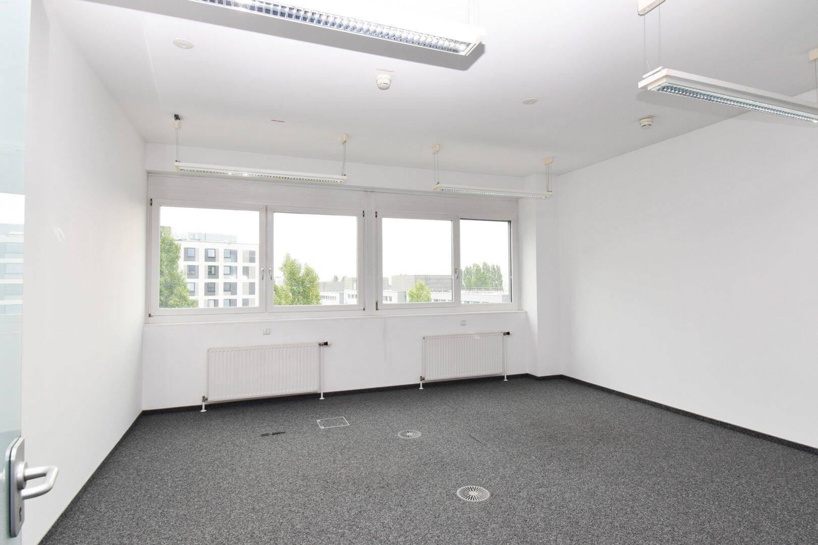 Leerer Büroraum mit Teppichboden, Heizkörpern unter den Fenstern und Blick auf die umliegenden Gebäude.