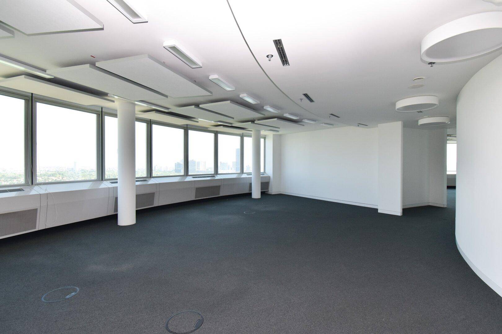 Großer, offener Bürobereich mit Teppichboden, Säulen und einer geschwungenen Wand, die einen Panoramablick bietet.
