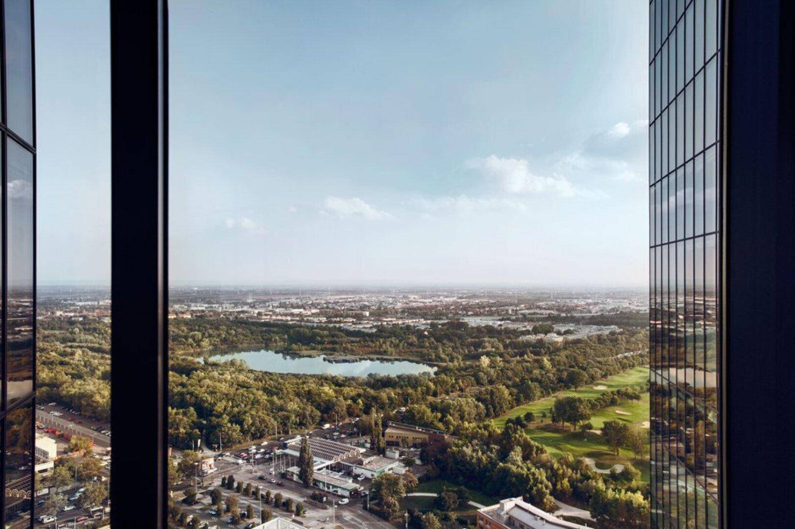Weitläufiger Blick aus einem Hochhausfenster auf eine grüne Landschaft mit See und Golfplatz unter klarem Himmel.