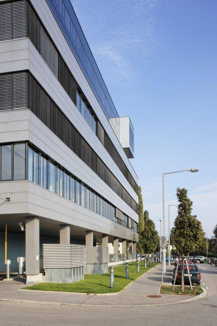 Seitenansicht eines modernen Bürogebäudes mit mehreren Etagen, großen Fenstern und einer gepflegten Grünfläche entlang der Straße.