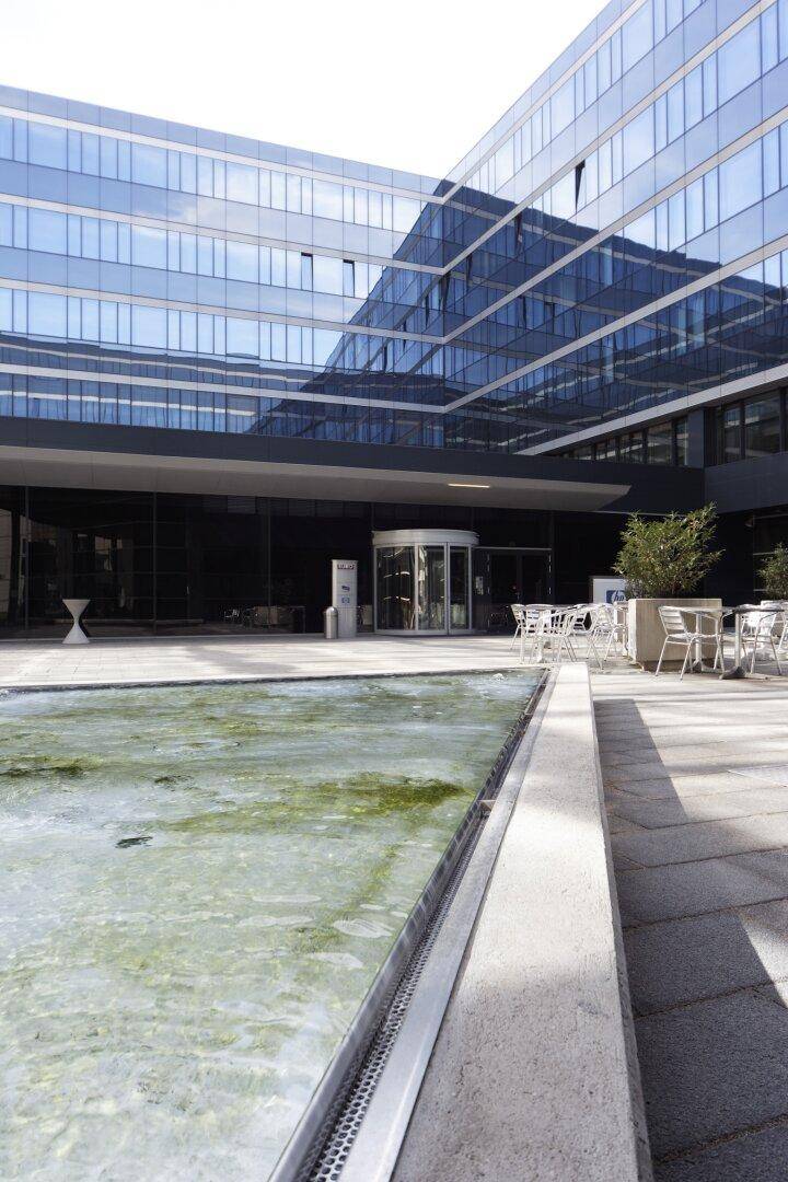 Innenhof eines Bürokomplexes mit einem Wasserbecken und Sitzgelegenheiten, umgeben von der modernen Glasfassade des Gebäudes.