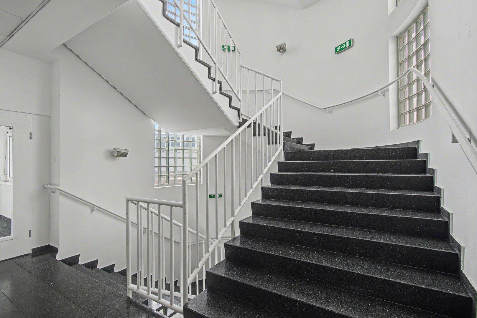 Treppenhaus mit dunklen Stufen, weißem Geländer und Glasbausteinen für natürliches Licht.