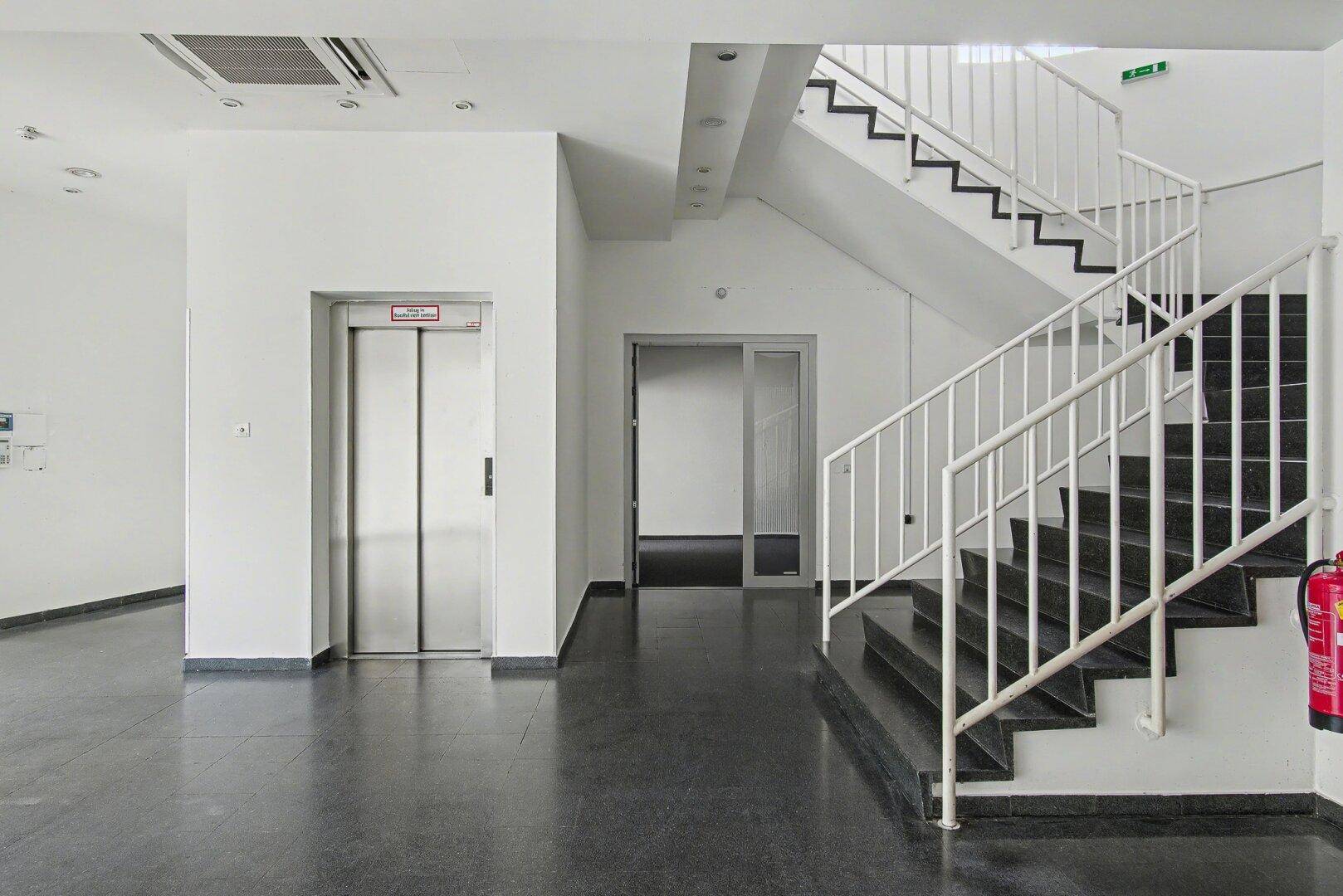 Heller Eingangsbereich mit Aufzug, Treppe und dunklem Steinboden.