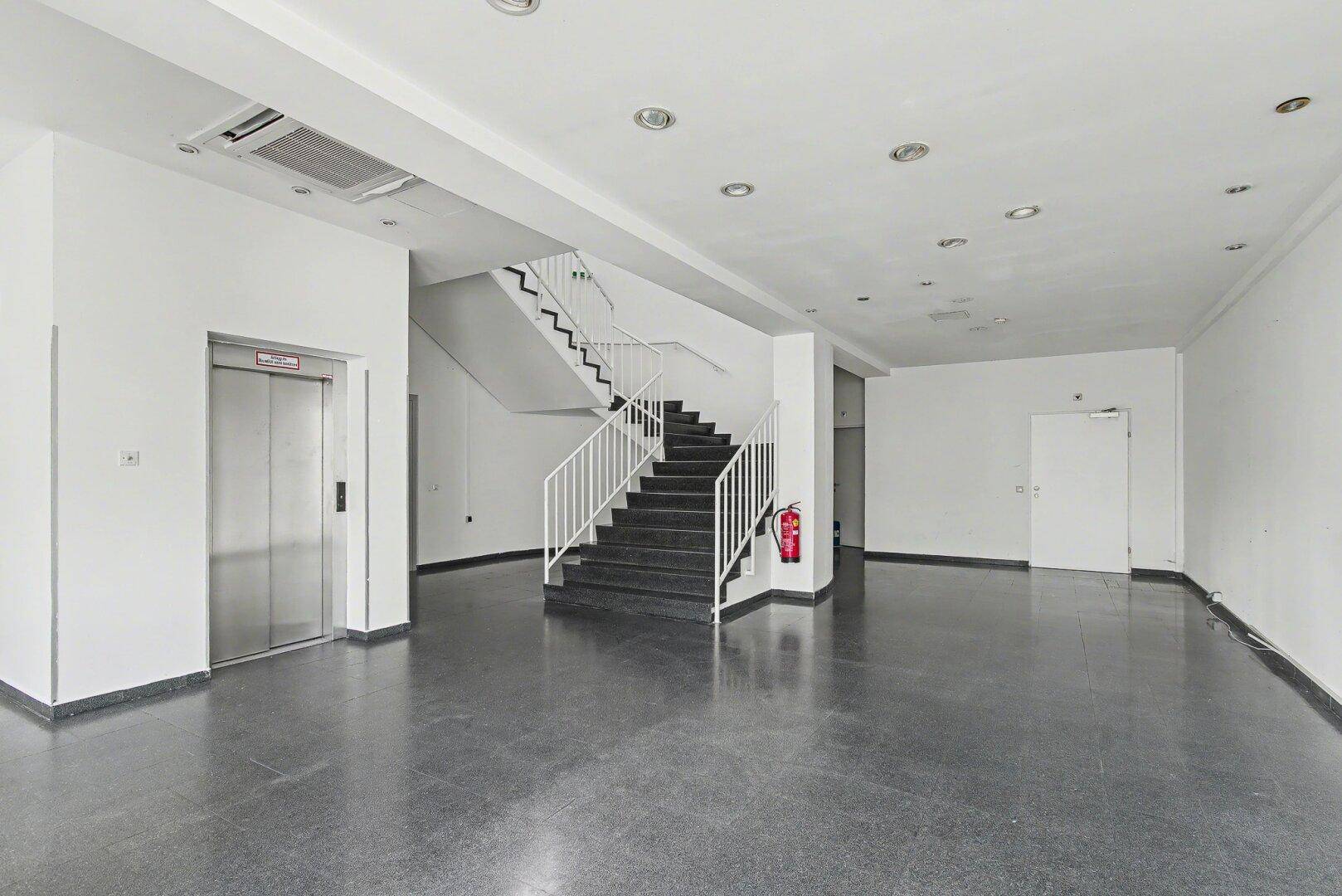 Weitläufiger Flurbereich mit Treppe, Aufzug und glänzendem dunklem Boden.