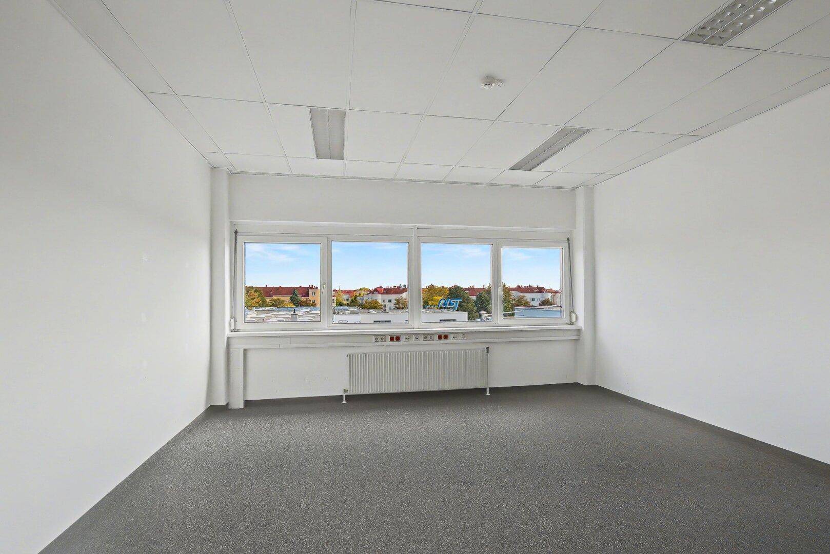 Helles Büro mit Teppichboden, großen Fenstern und Blick auf die Dächer der Stadt.
