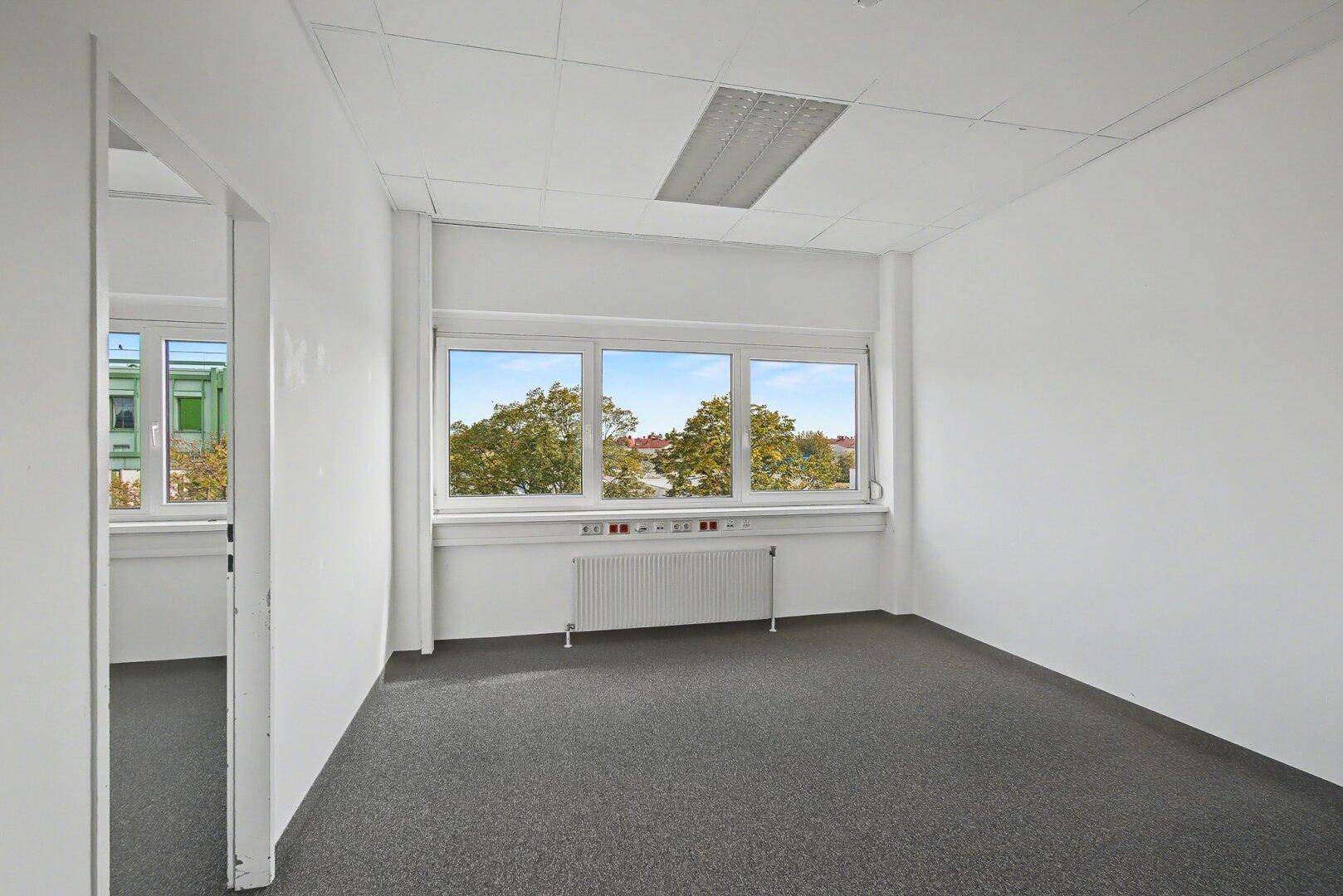Leeres Büro mit Teppichboden, großen Fenstern und Blick auf die umliegenden Gebäude.