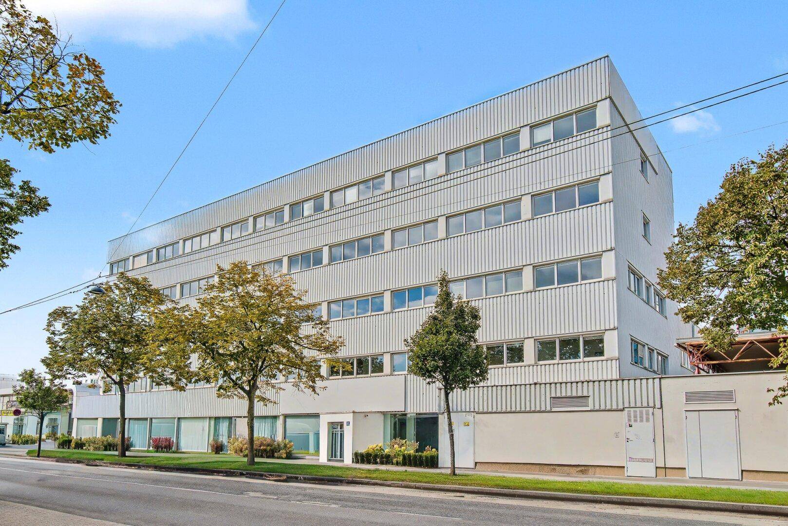 Modernes Bürogebäude mit mehreren Etagen und vielen Fenstern an einer Straße mit Bäumen.