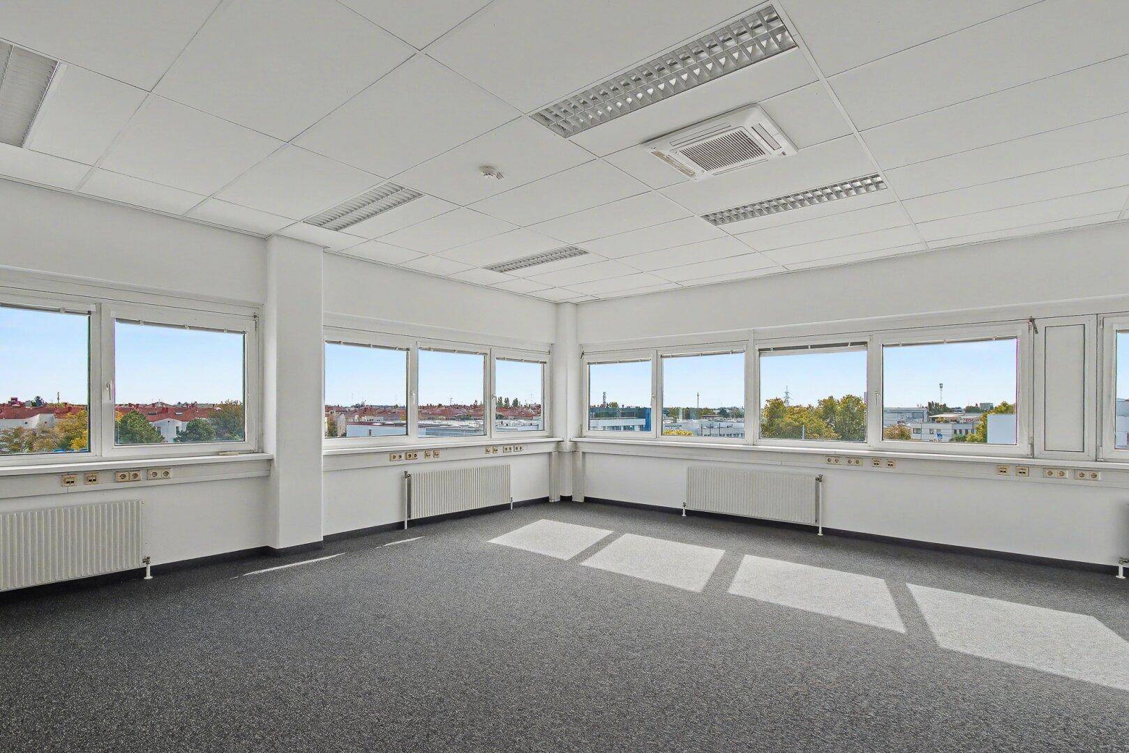 Geräumiges, leeres Büro mit Teppichboden, vielen Fenstern und Blick auf die Stadt.