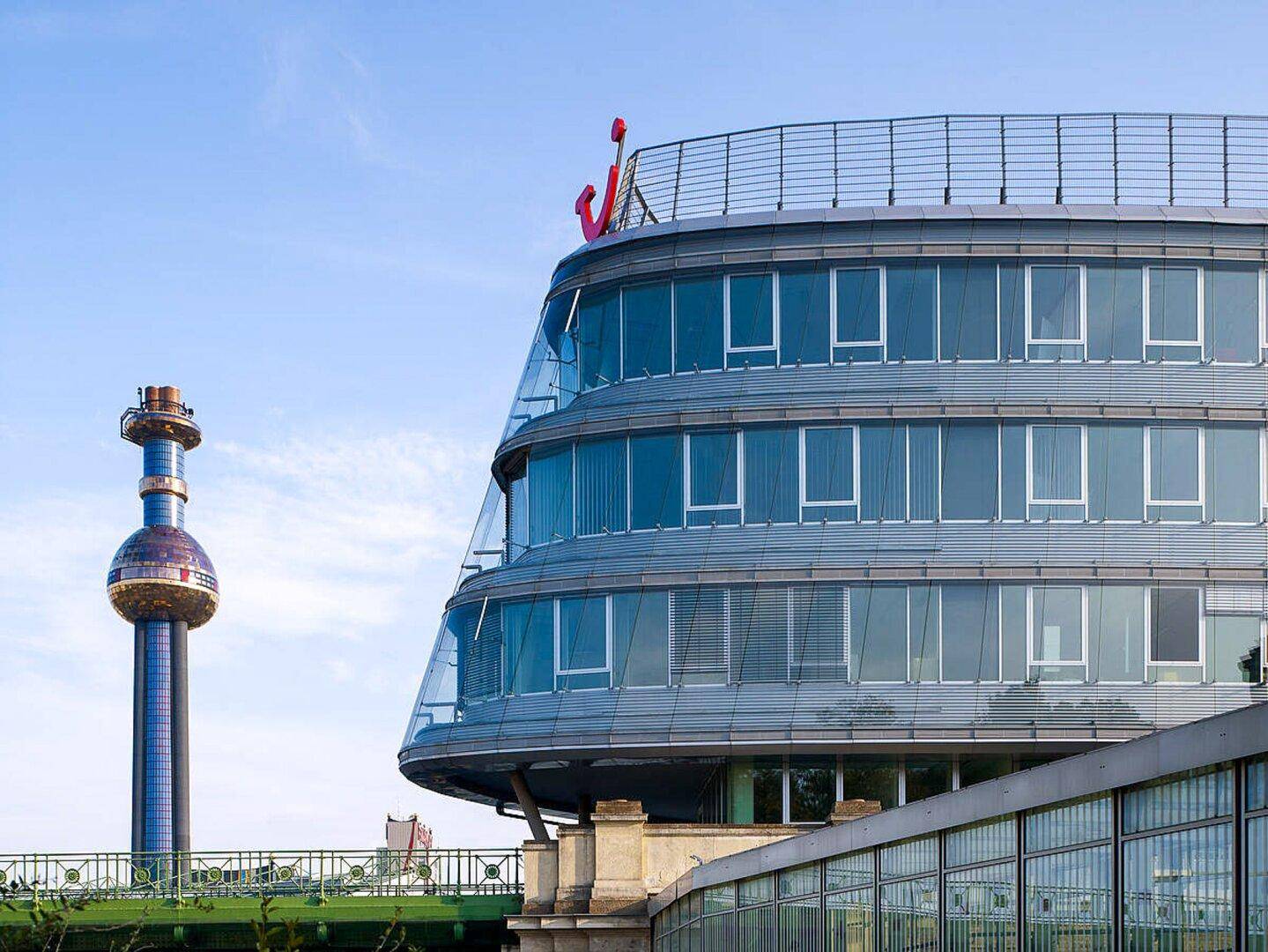 Gebogenes modernes Bürogebäude mit Glasfassade und einem markanten Turm im Hintergrund unter blauem Himmel.