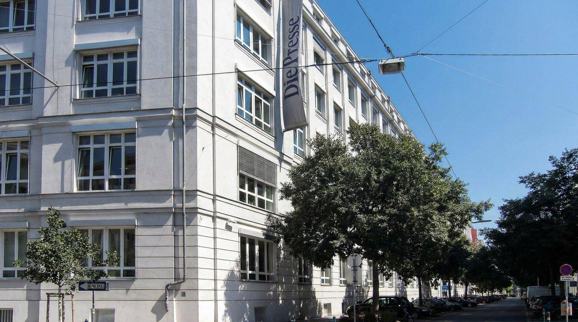 Ecke eines weißen Bürogebäudes mit großem Banner und Bäumen entlang der Straße.