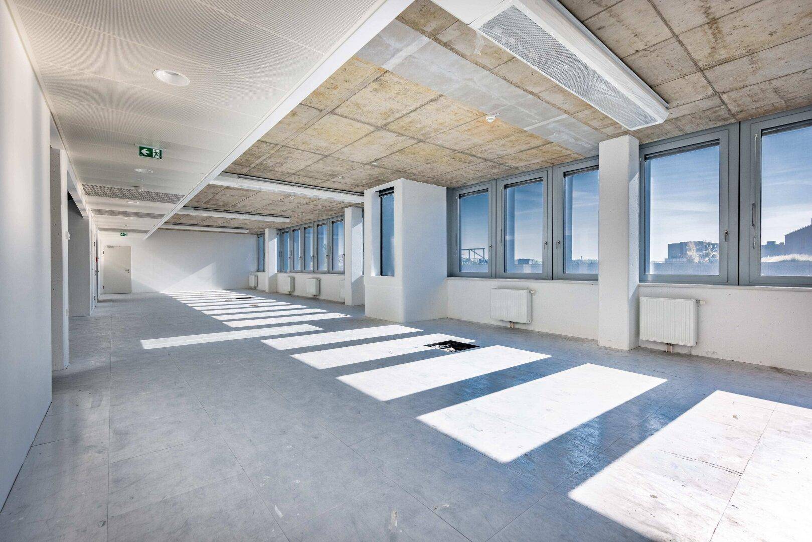 Heller Bürogang mit Fliesenboden, Beton-Decke und langen Sonnenstrahlen durch die Fenster.
