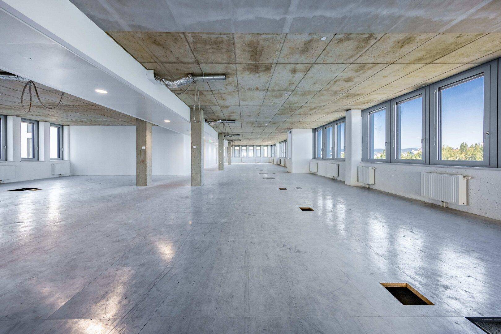 Großer, leerer Büroraum mit Fliesenboden, Beton-Decke und vielen Fenstern.
