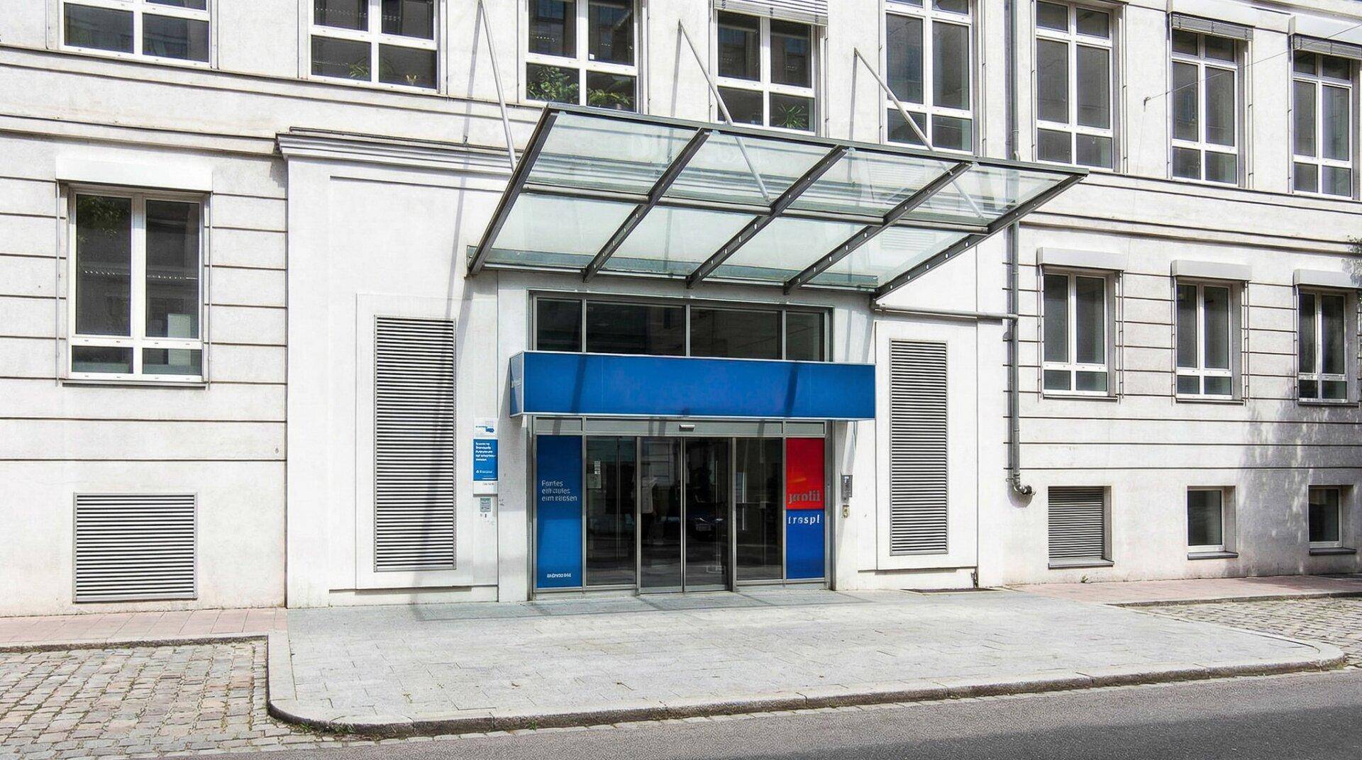 Modernes Gebäude mit Glasvordach und automatischen Türen am Haupteingang, umgeben von einer sauberen Gehwegfläche.