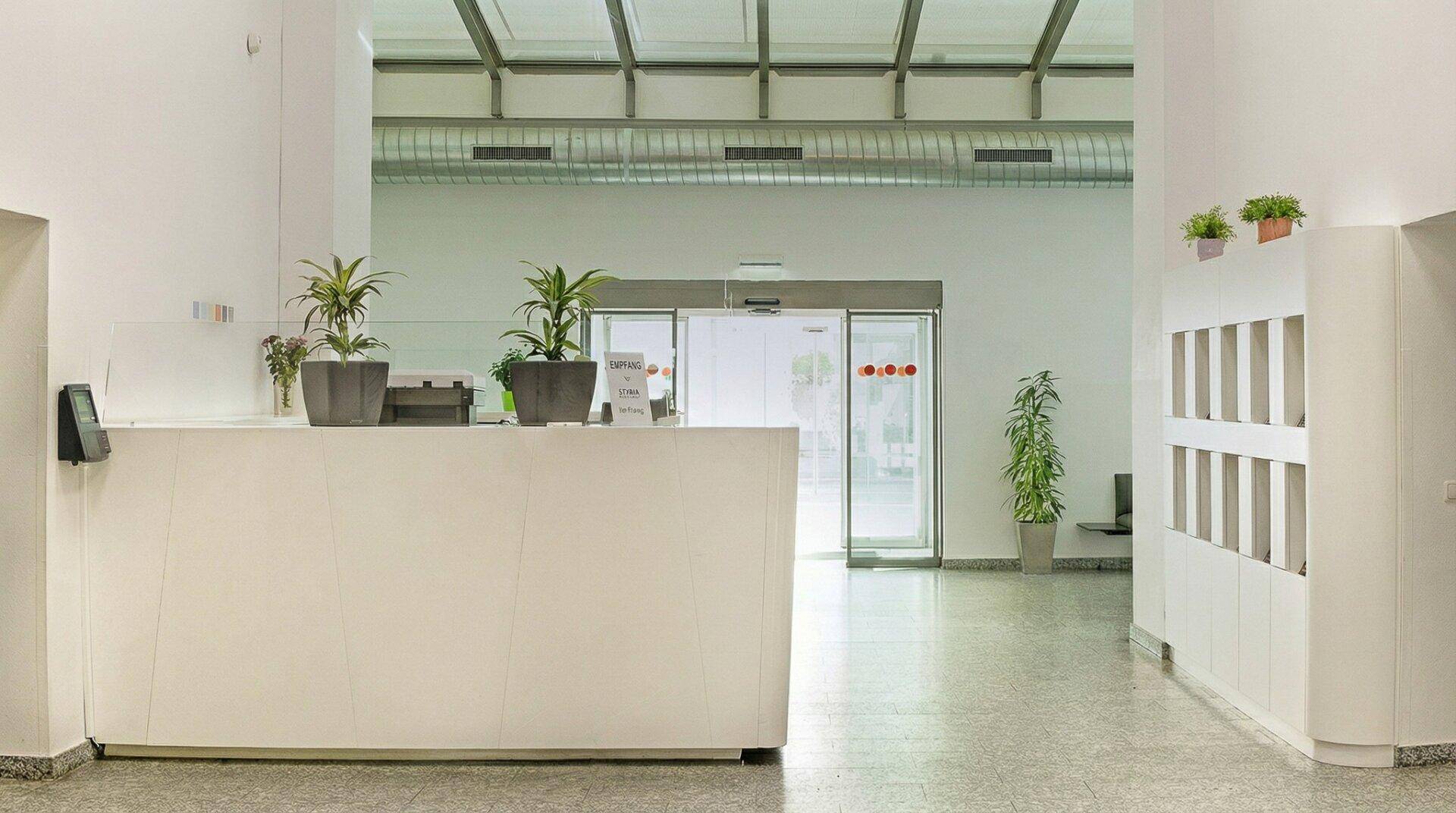 Heller Empfangsbereich mit moderner Rezeptionstheke und Pflanzen, führt zu automatischen Glastüren und hat sichtbare Lüftungskanäle.