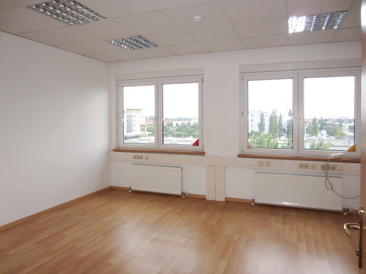 Helles Büro mit Laminatboden, großen Fenstern und Blick auf Bäume und Gebäude, bietet viel Tageslicht.