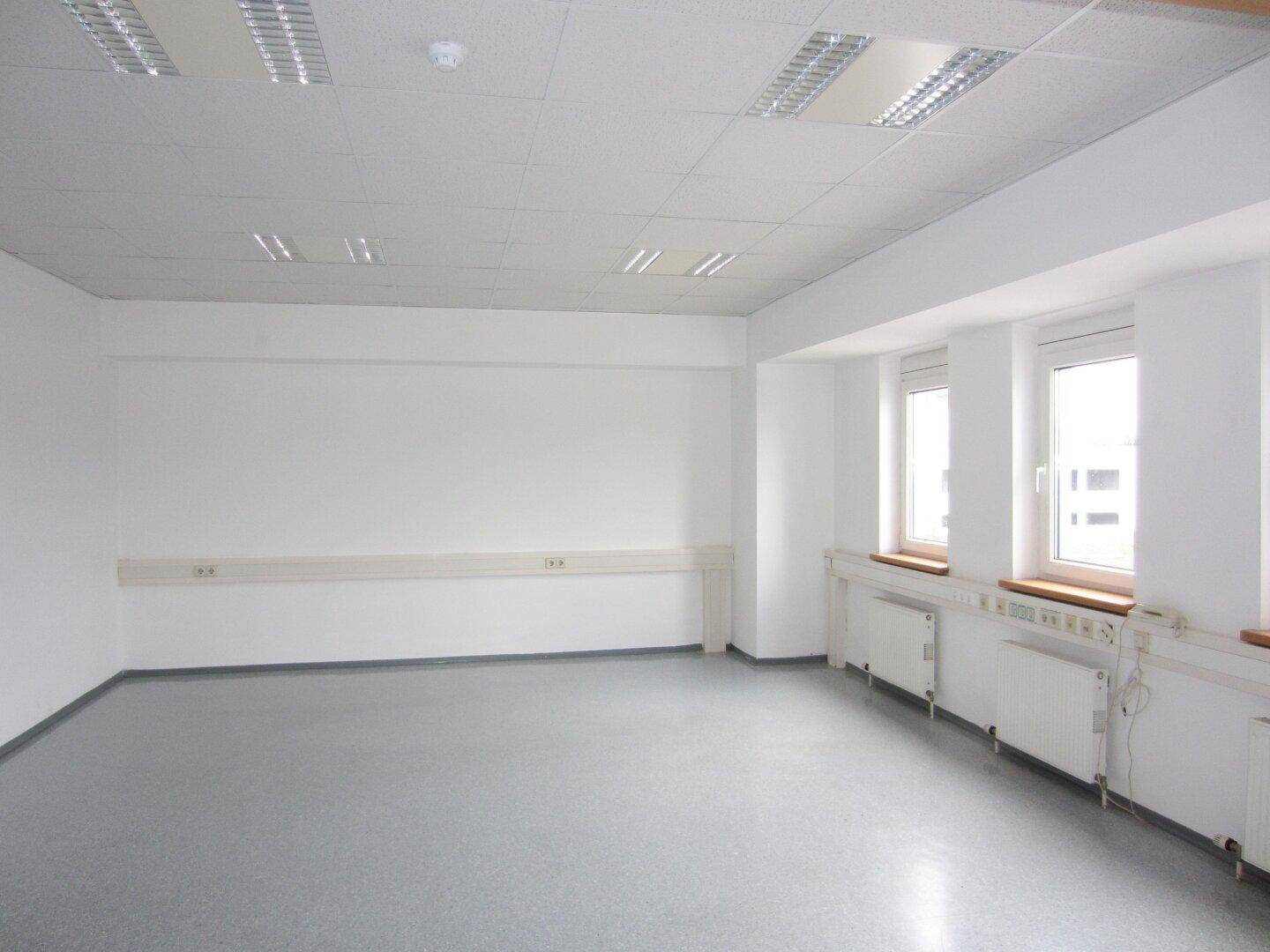 Helles, leeres Büro mit grauem Vinylboden, großen Fenstern und Heizkörpern unter den Fensterbänken.