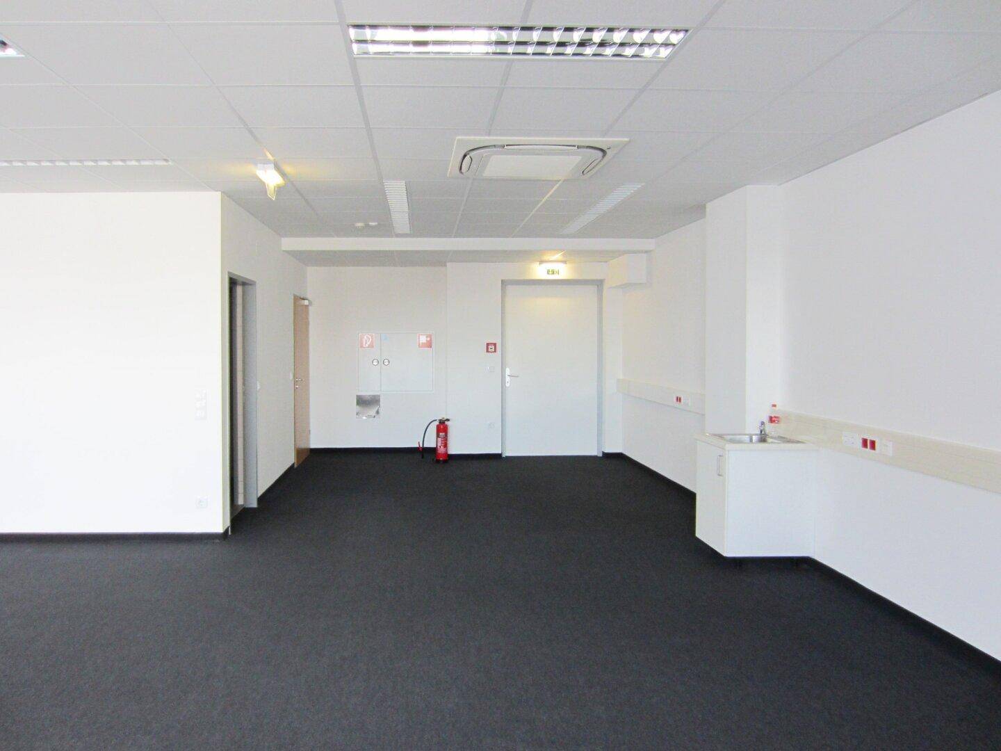 Geräumiges Büro mit dunklem Teppichboden, Brandschutzvorrichtungen und einer kleinen Spüle in der Ecke.