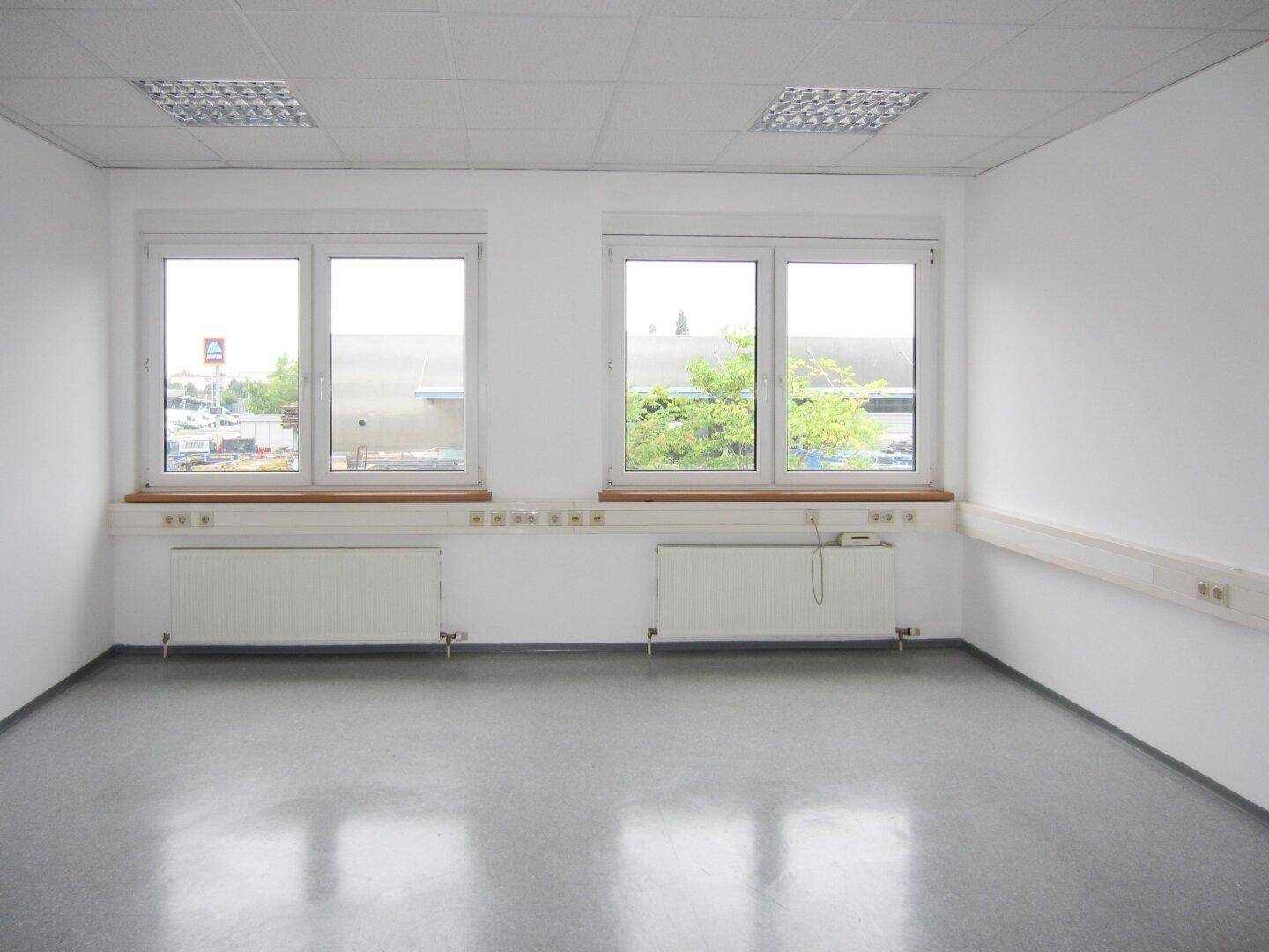 Leeres Büro mit grauem Vinylboden, zwei großen Fenstern und Blick auf die umliegende Bebauung.
