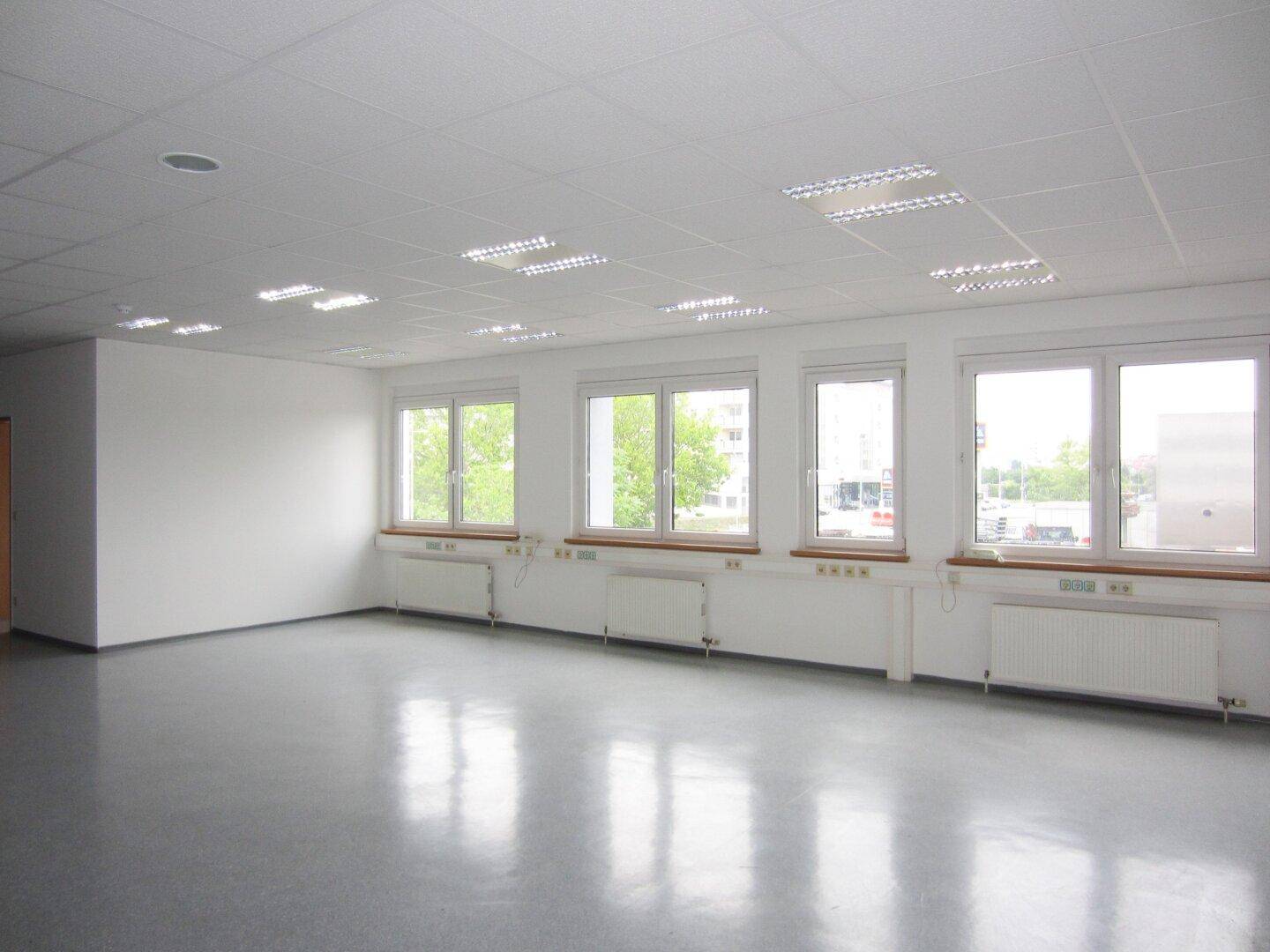 Helles, leeres Büro mit grauem Vinylboden, vielen Fenstern und Blick auf die Umgebung, bietet viel Tageslicht.