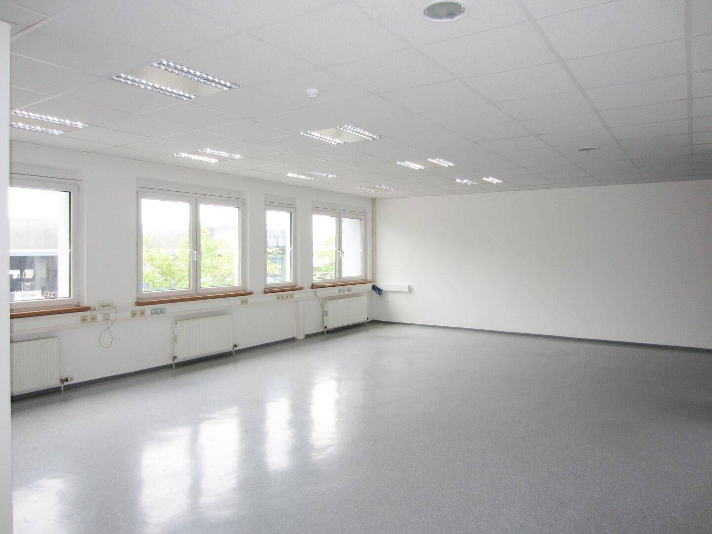 Großes, helles Büro mit grauem Vinylboden, vielen Fenstern und Blick auf die Umgebung, ideal für Teamarbeit.