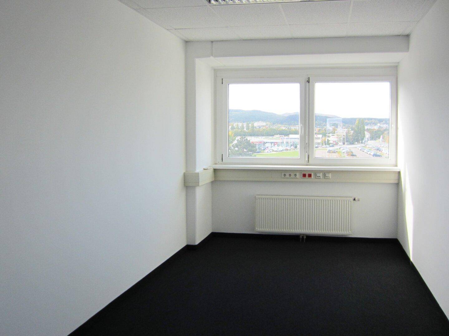 Kleines, helles Büro mit dunklem Teppichboden, großem Fenster und Blick auf die Stadt, ideal für Einzelbüros.