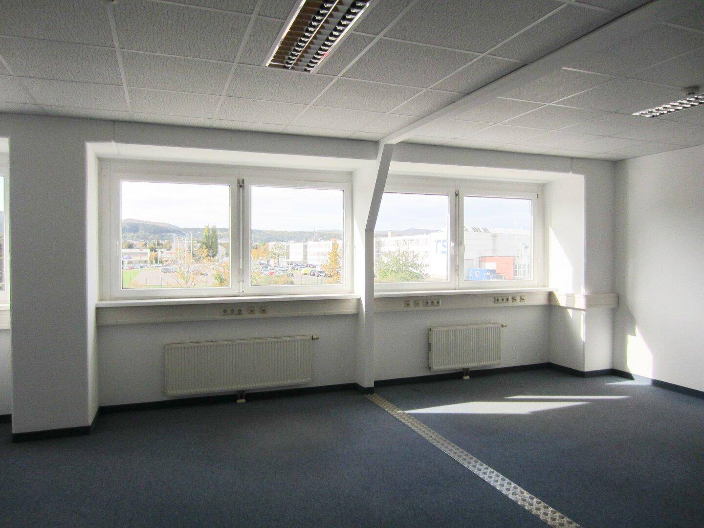 Großes, helles Büro mit blauem Teppichboden, vielen Fenstern und Blick auf die Umgebung, ideal für kreative Arbeit.