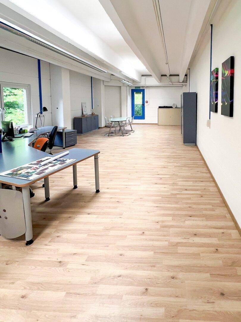 Langer, offener Bürobereich mit hellem Holzboden, mehreren Arbeitsplätzen und einem Besprechungsbereich.