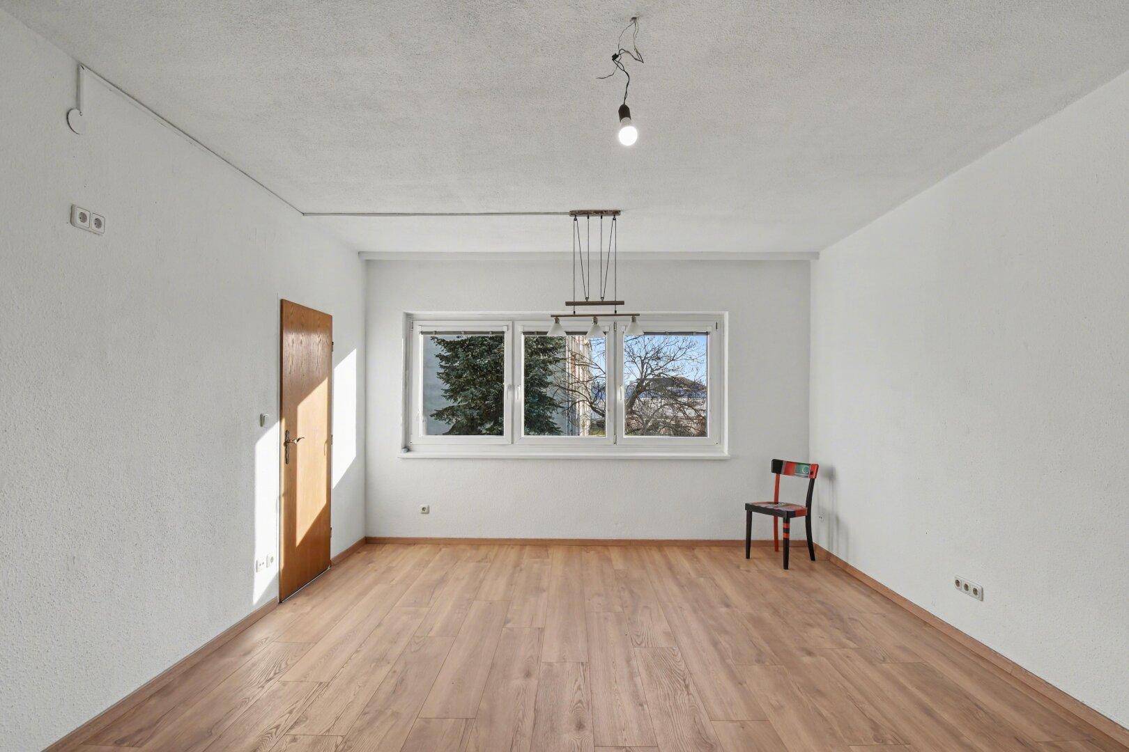Geräumiger, leerer Raum mit Laminatboden und einem großen Fenster, das den Blick nach draußen freigibt.