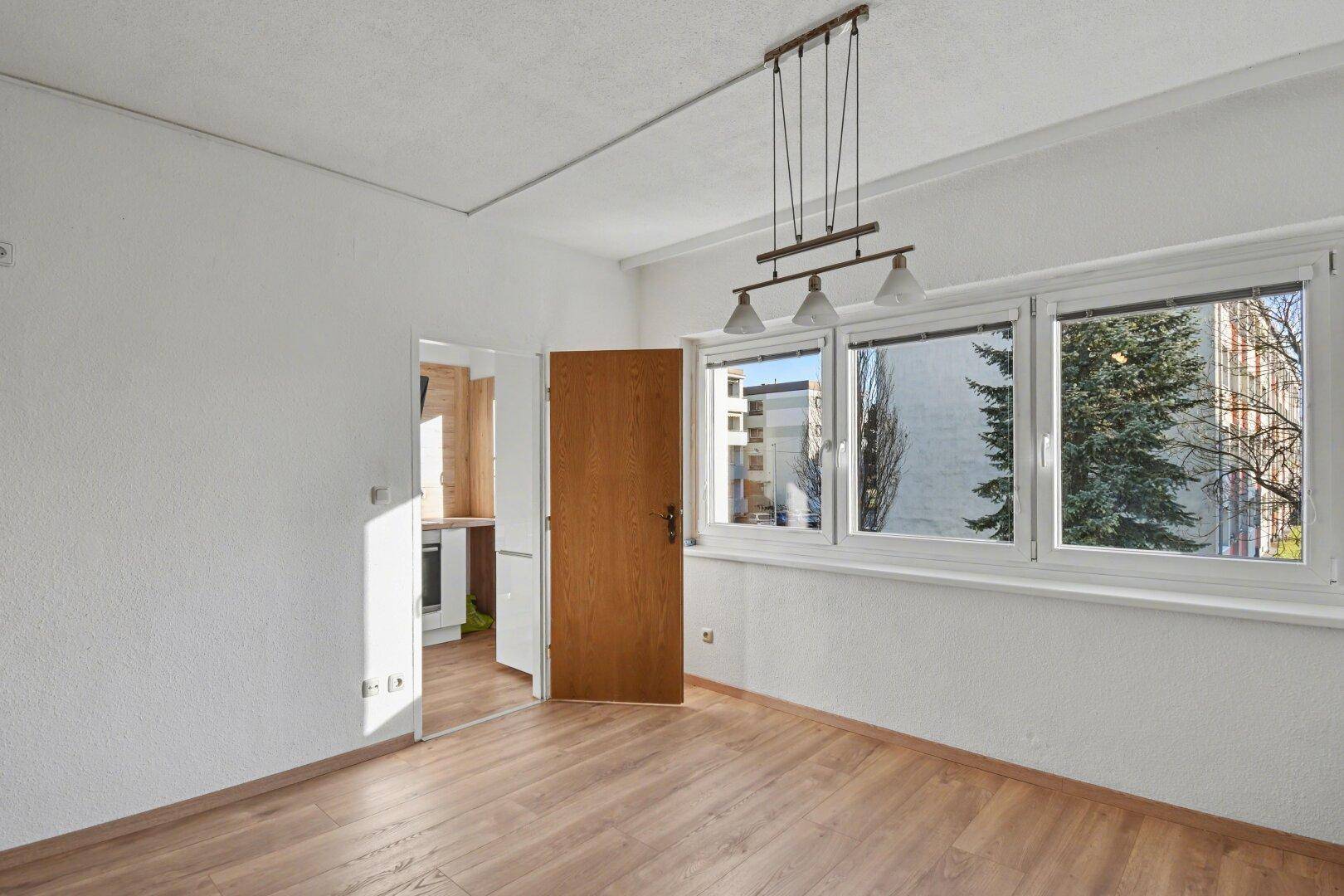 Heller Wohnbereich mit Laminatboden und Zugang zu einer angrenzenden Küche durch eine offene Tür.