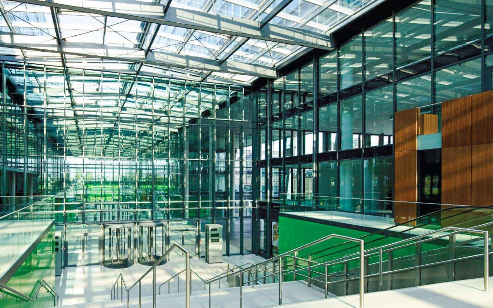 Helle, moderne Lobby mit Glasfassade, Treppenaufgang und viel Grünfläche im Hintergrund.