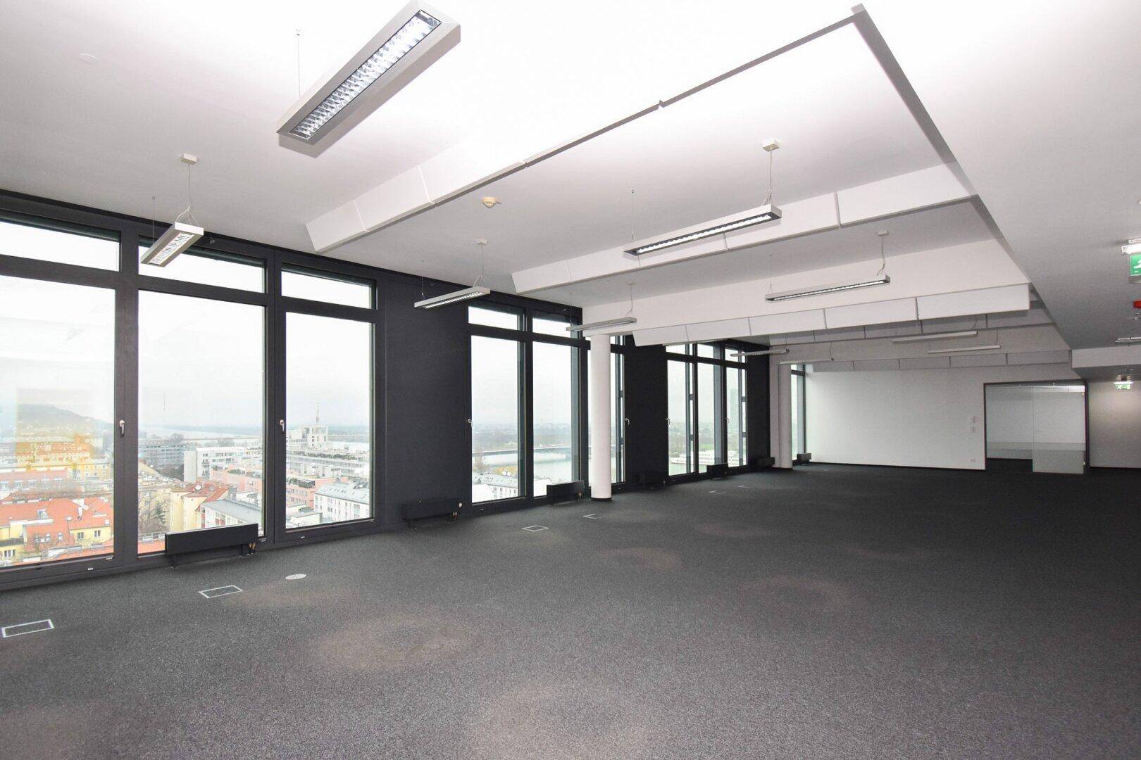 Geräumiges, leeres Büro mit großen Fenstern, Teppichboden und Panoramablick auf die Stadt.