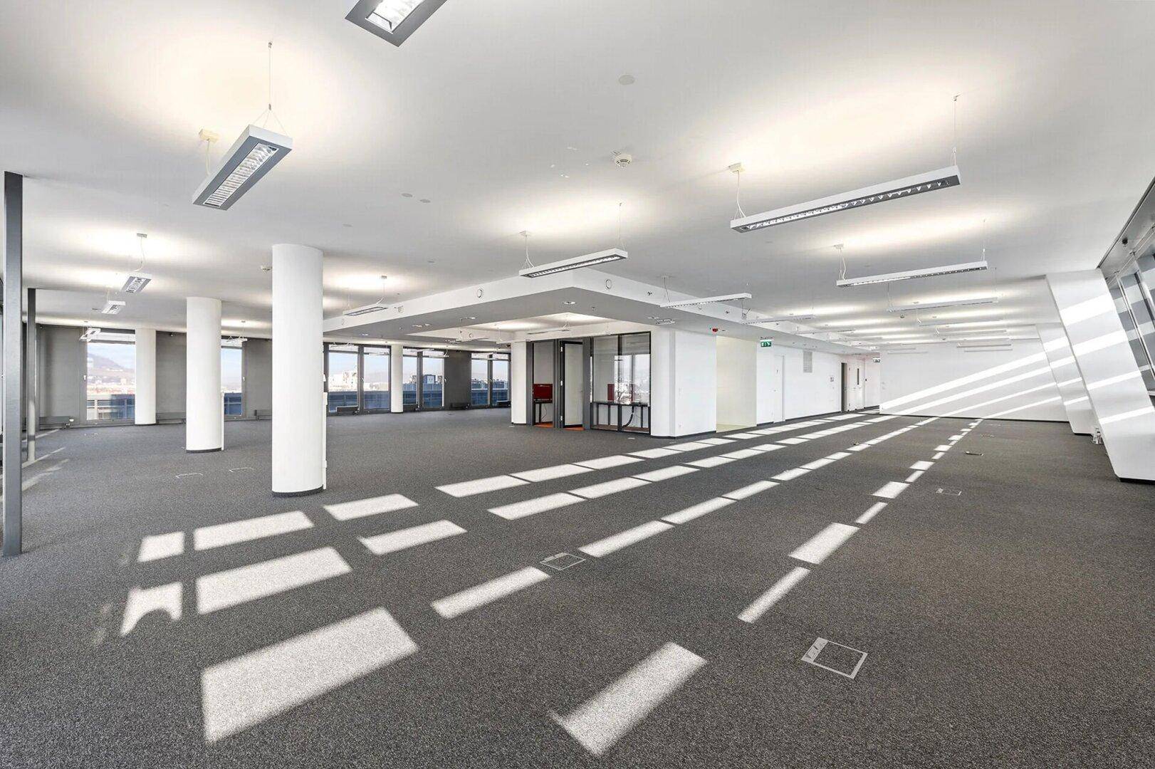 Weitläufiger, offener Bürobereich mit Teppichboden, Säulen und großen Fenstern, die einen Panoramablick auf die Stadt bieten.