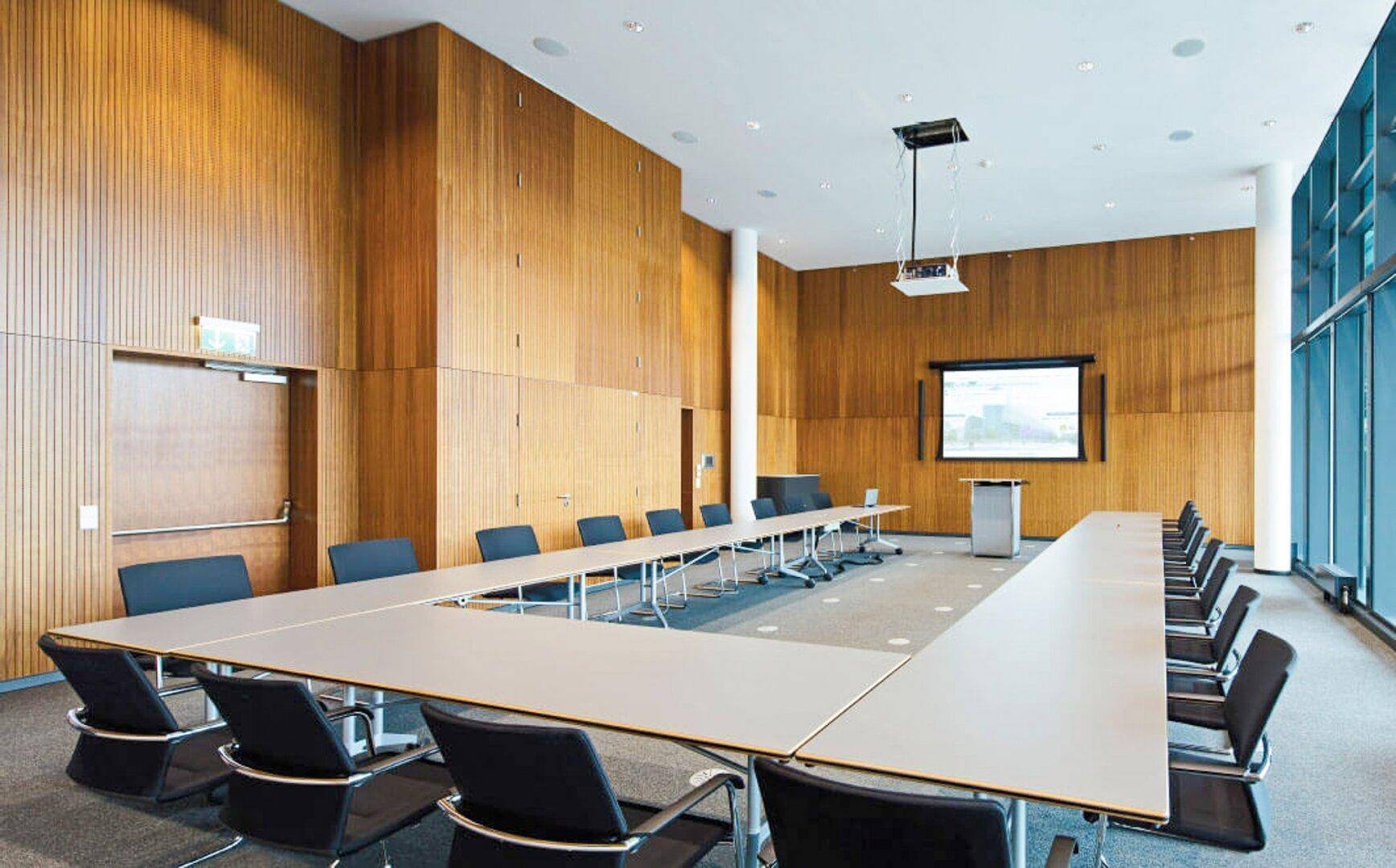 Großer Konferenzraum mit Holzwänden, Teppichboden, Projektor und U-förmiger Tischordnung, ideal für professionelle Besprechungen.